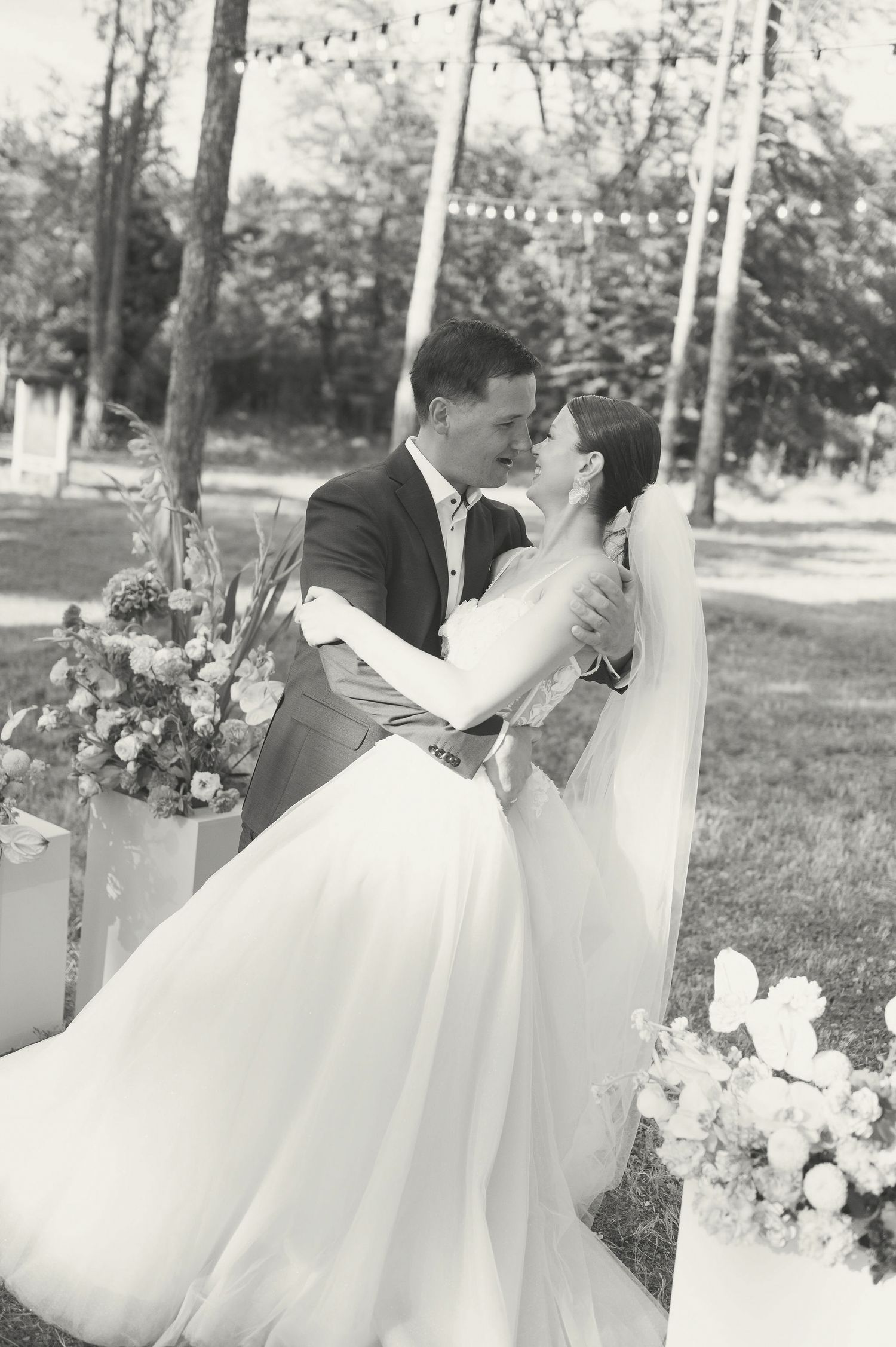 A black and white wedding portrait showing a romantic embrace under string lights and trees.