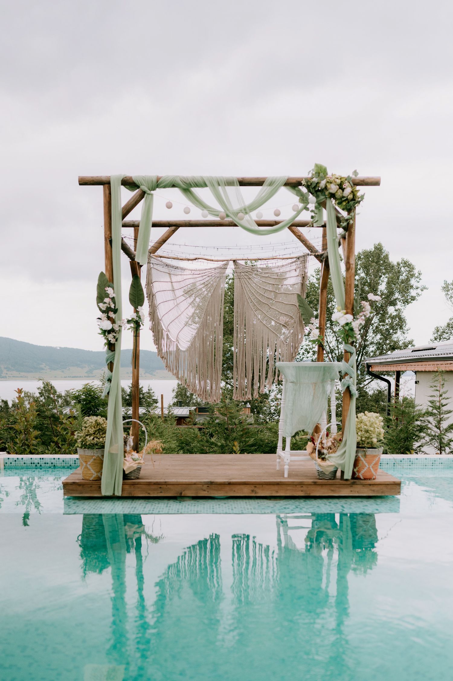 Elegant wedding arch with flowing macrame panels and greenery beside a swimming pool.