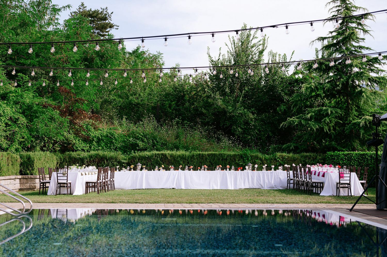 Long outdoor dining tables set up under string lights with a swimming pool in the background.