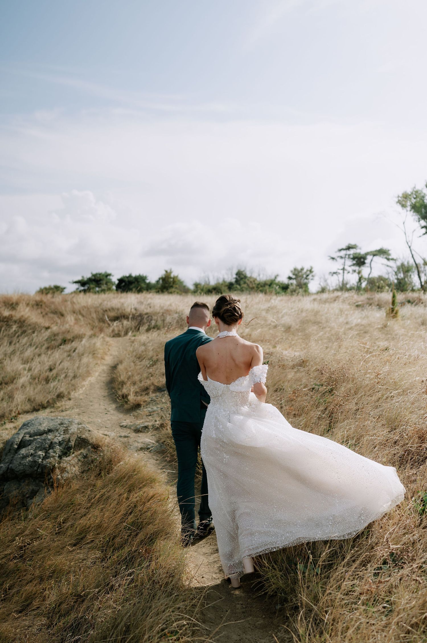 Wedding couple walks through golden grass field with flowing white dress catching the wind.