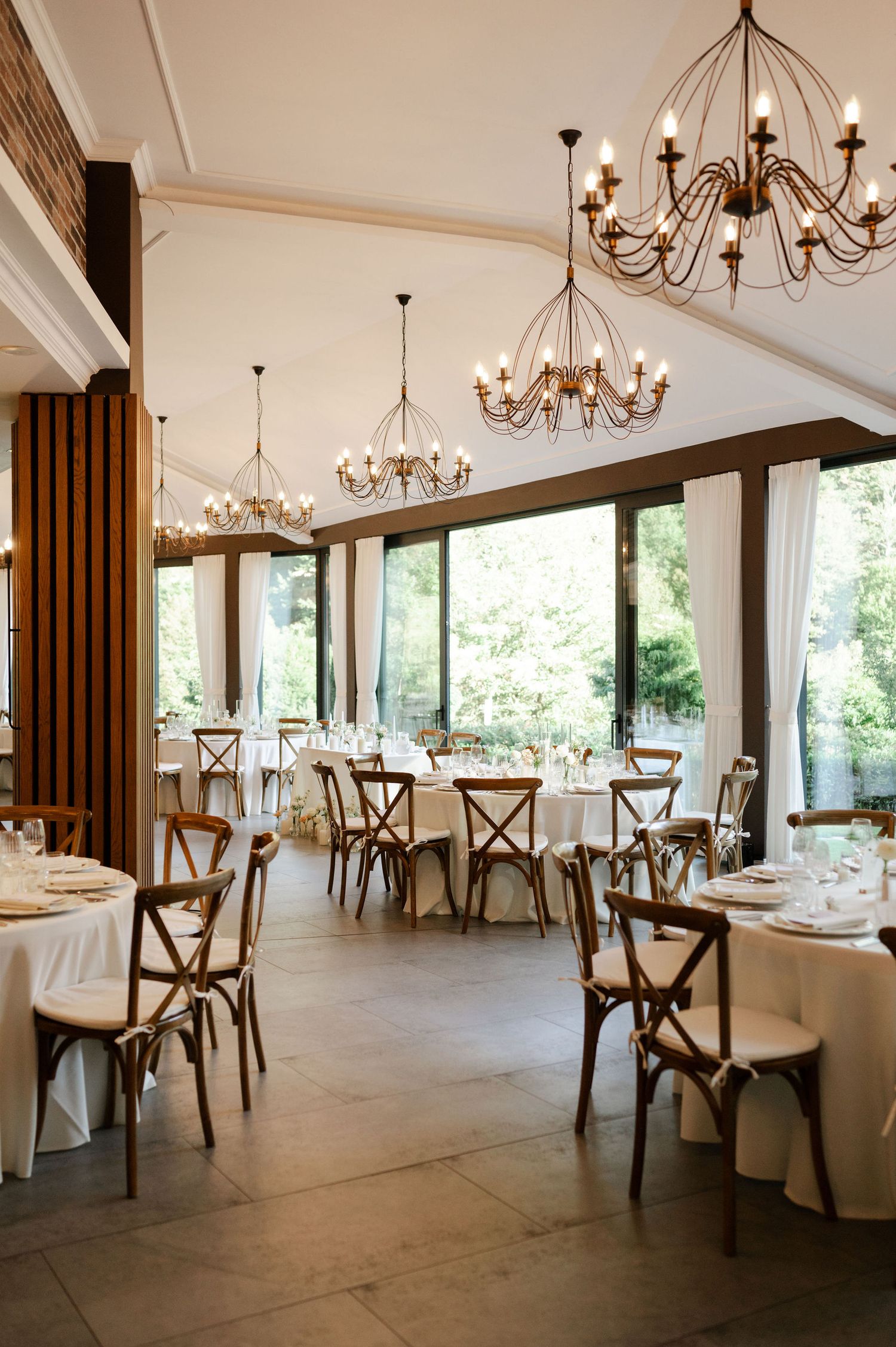 Elegant wedding venue interior with chandeliers, white tablecloths and cross-back chairs.