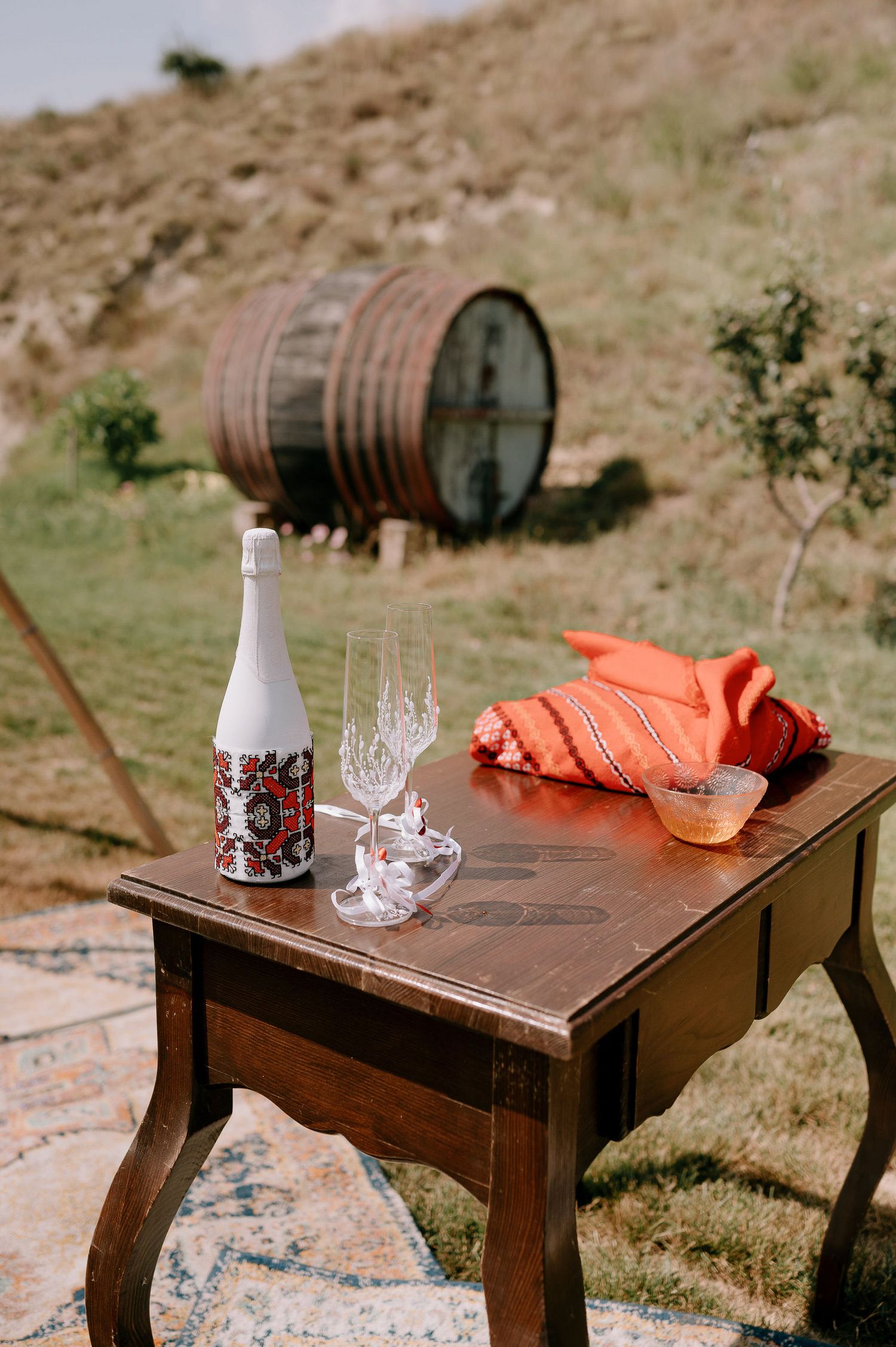 Vintage table with wine bottle and glasses set against rustic wine barrel in countryside.