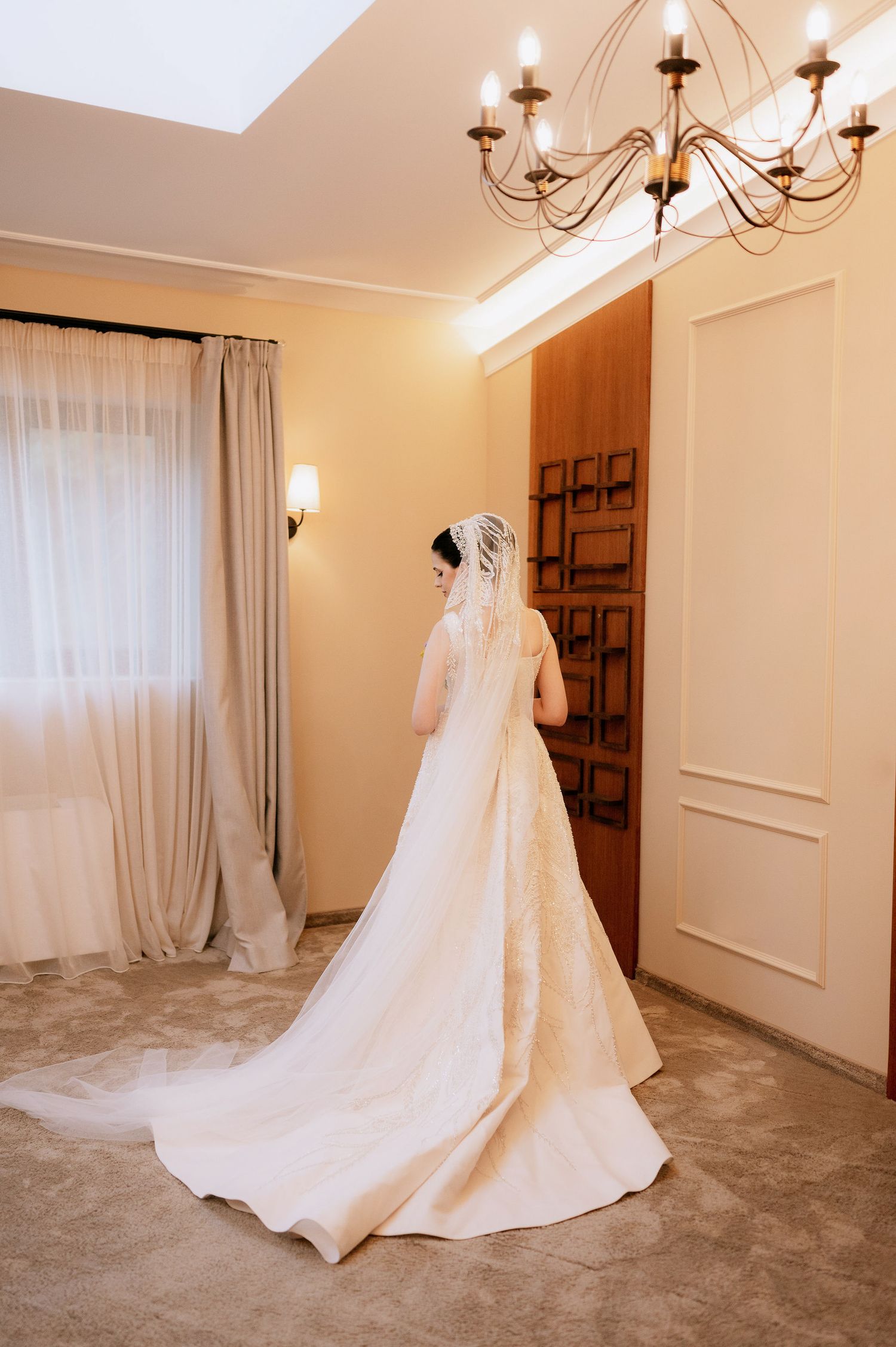 An elegant white wedding gown with a flowing train displayed in a room with chandelier lighting.