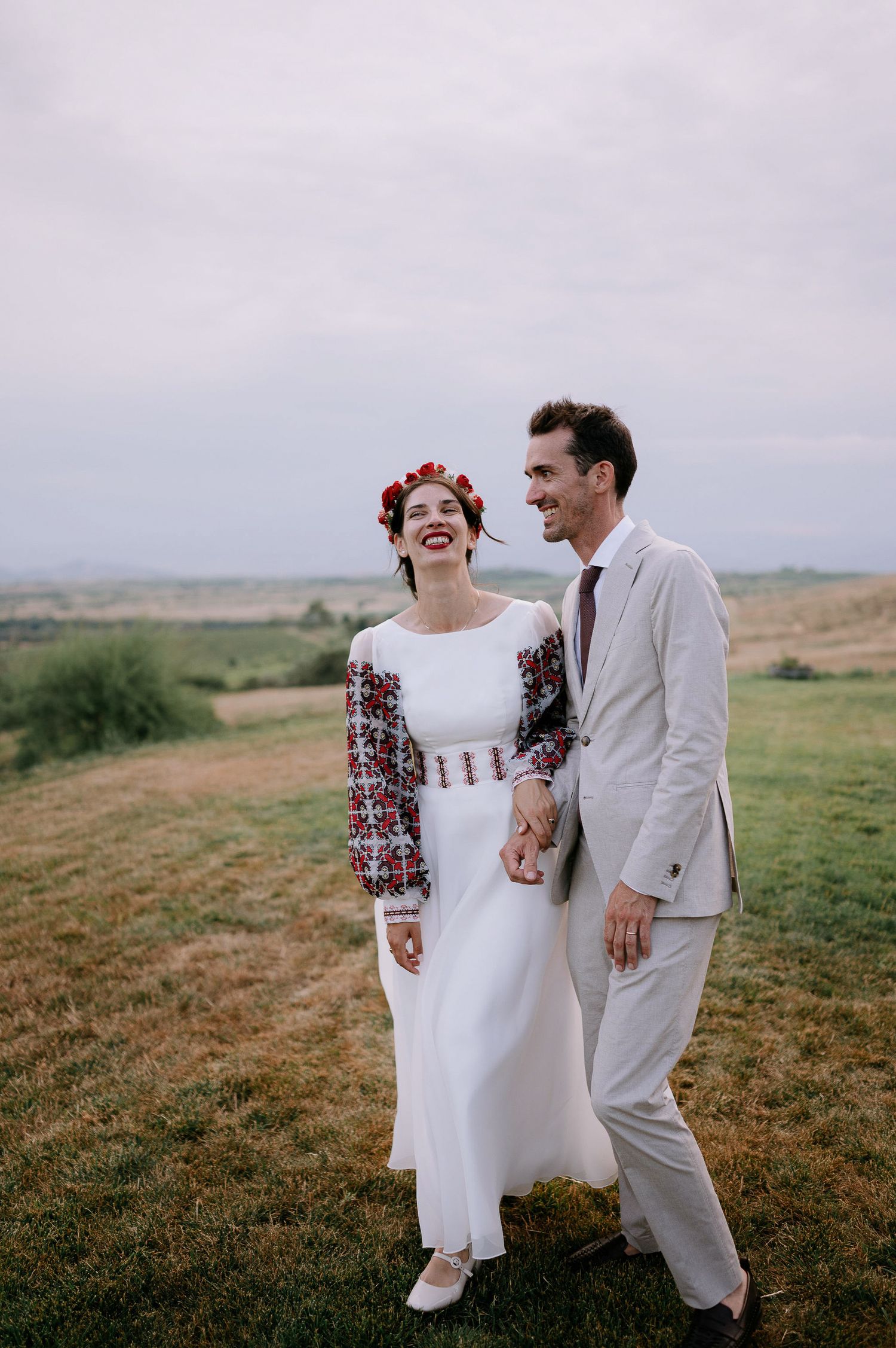 Newlyweds in folk-inspired dress dance and laugh together in a hilltop meadow.