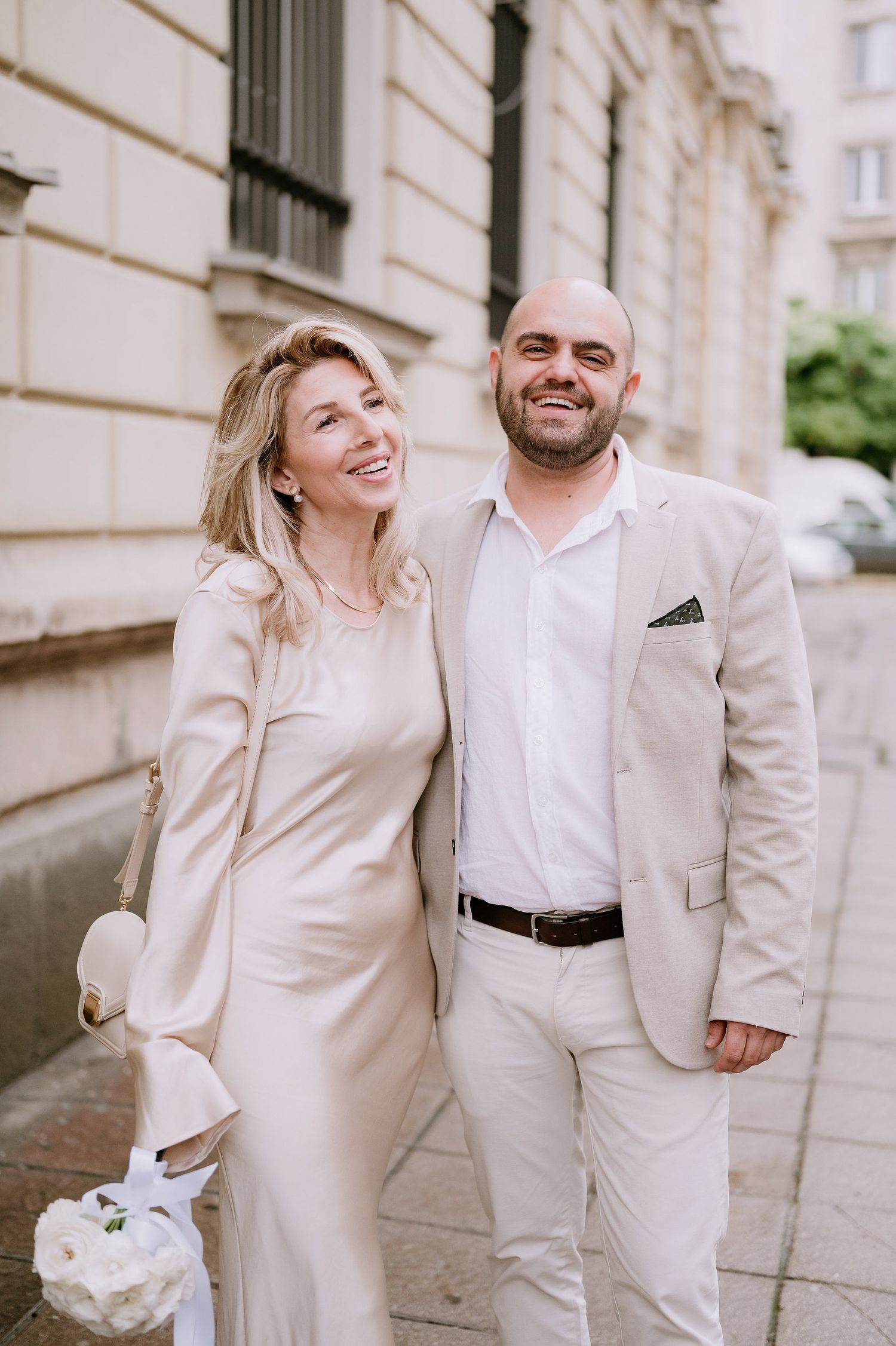 Joyful couple in light colored formal wear sharing a candid moment on the sidewalk.