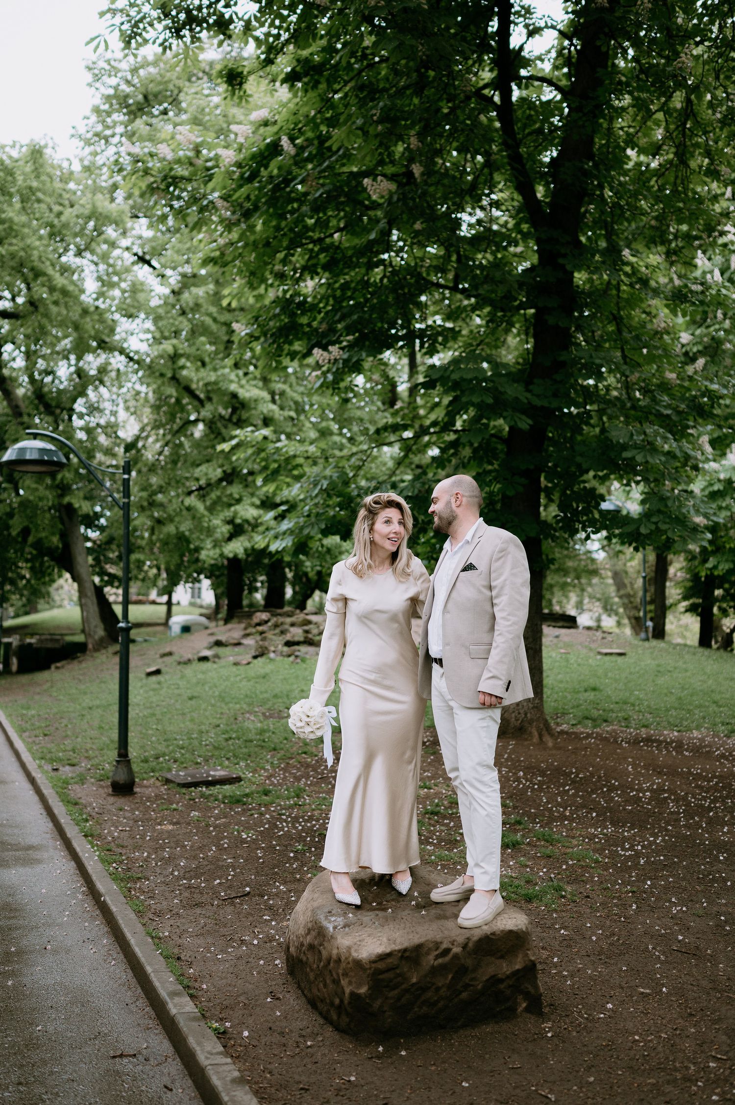 A couple in coordinated cream outfits poses near a large boulder in a park landscape.