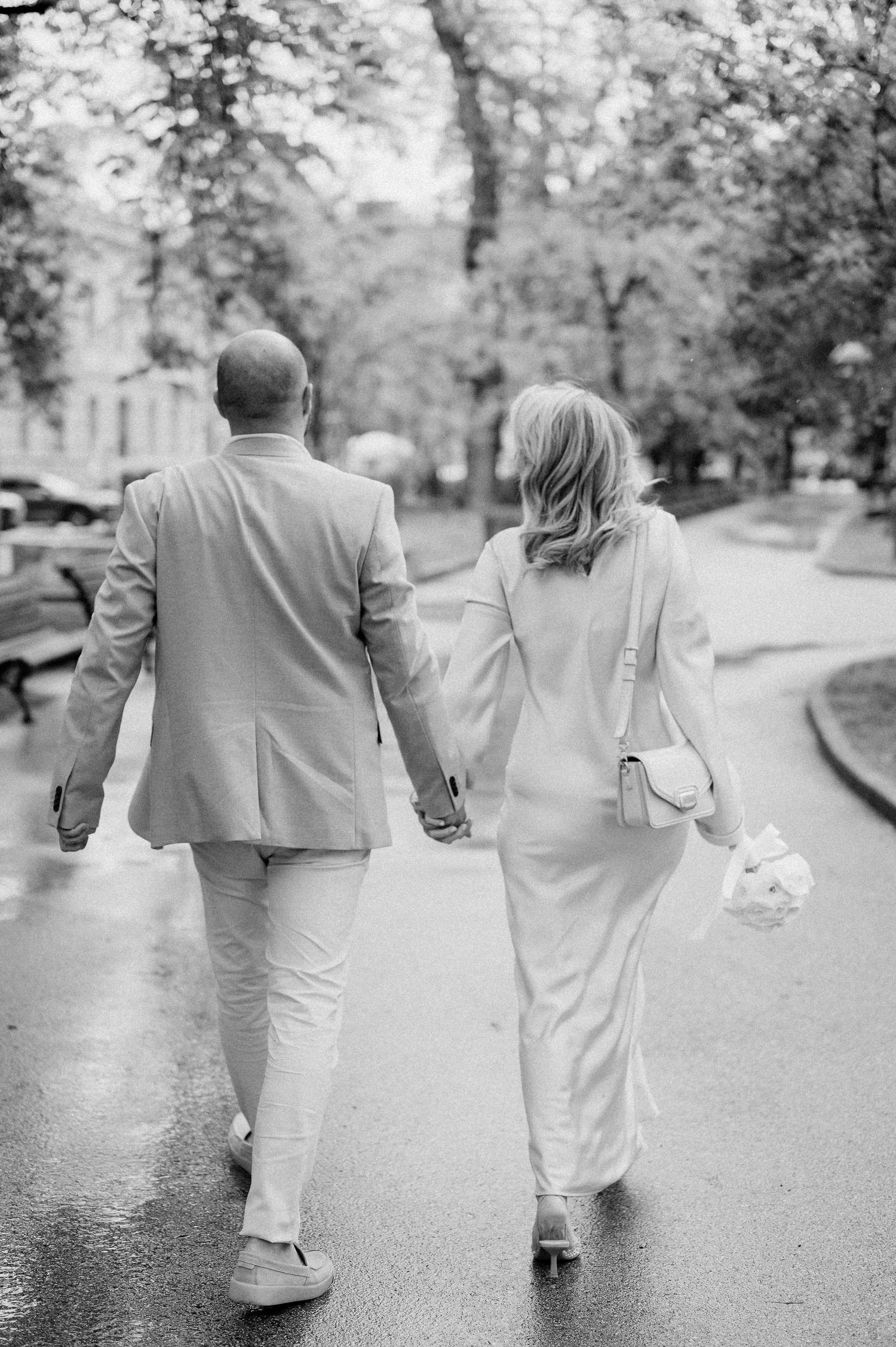 A couple in light-colored outfits walking together on a wet pathway through a park.