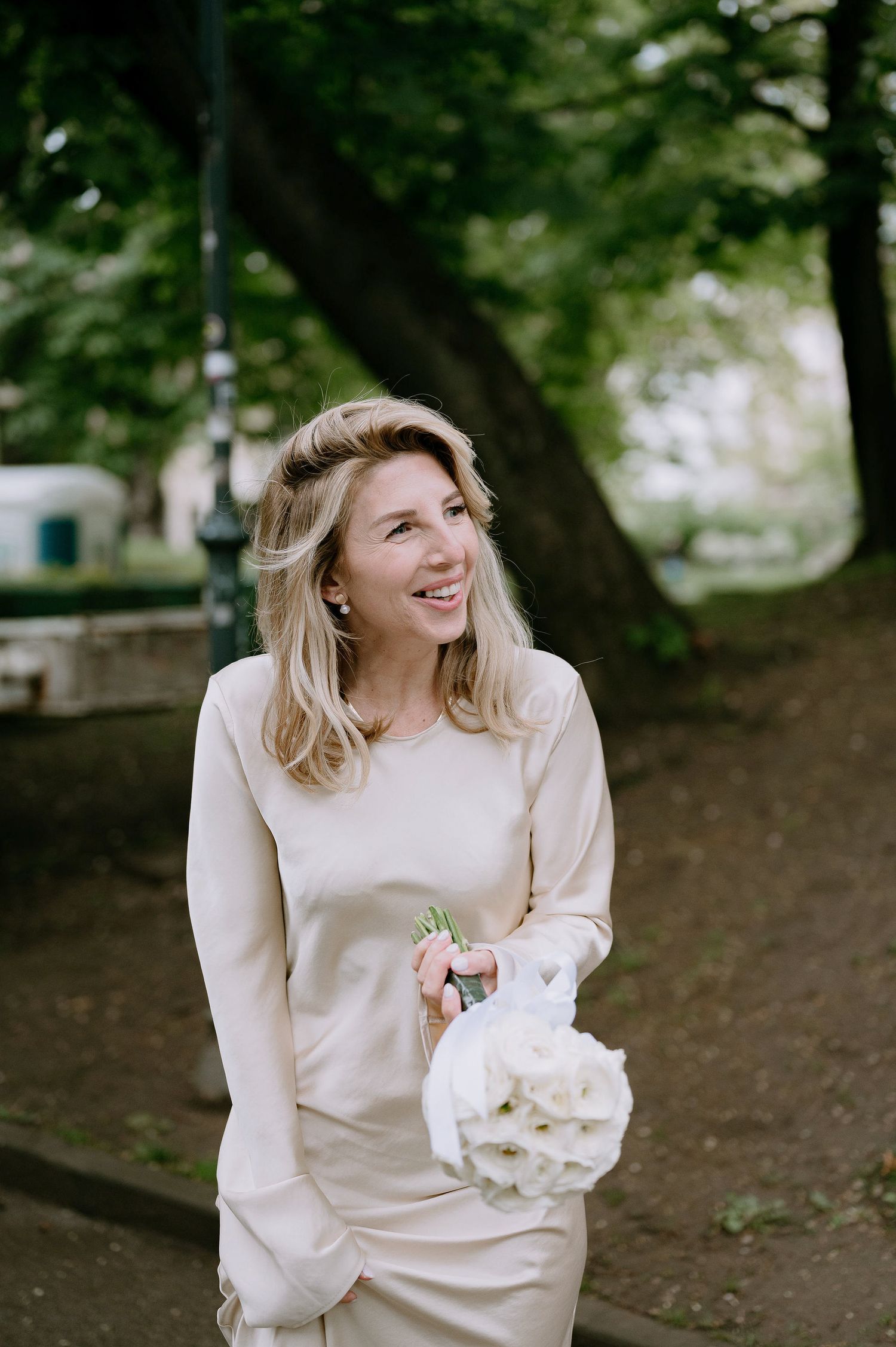 A person in cream-colored clothing smiles while holding a white bouquet in a park setting.