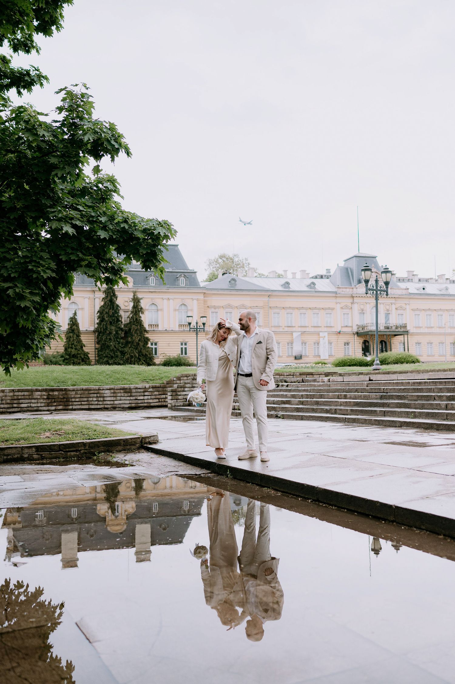 Reflections in water mirror two figures standing near stone steps at a grand estate.