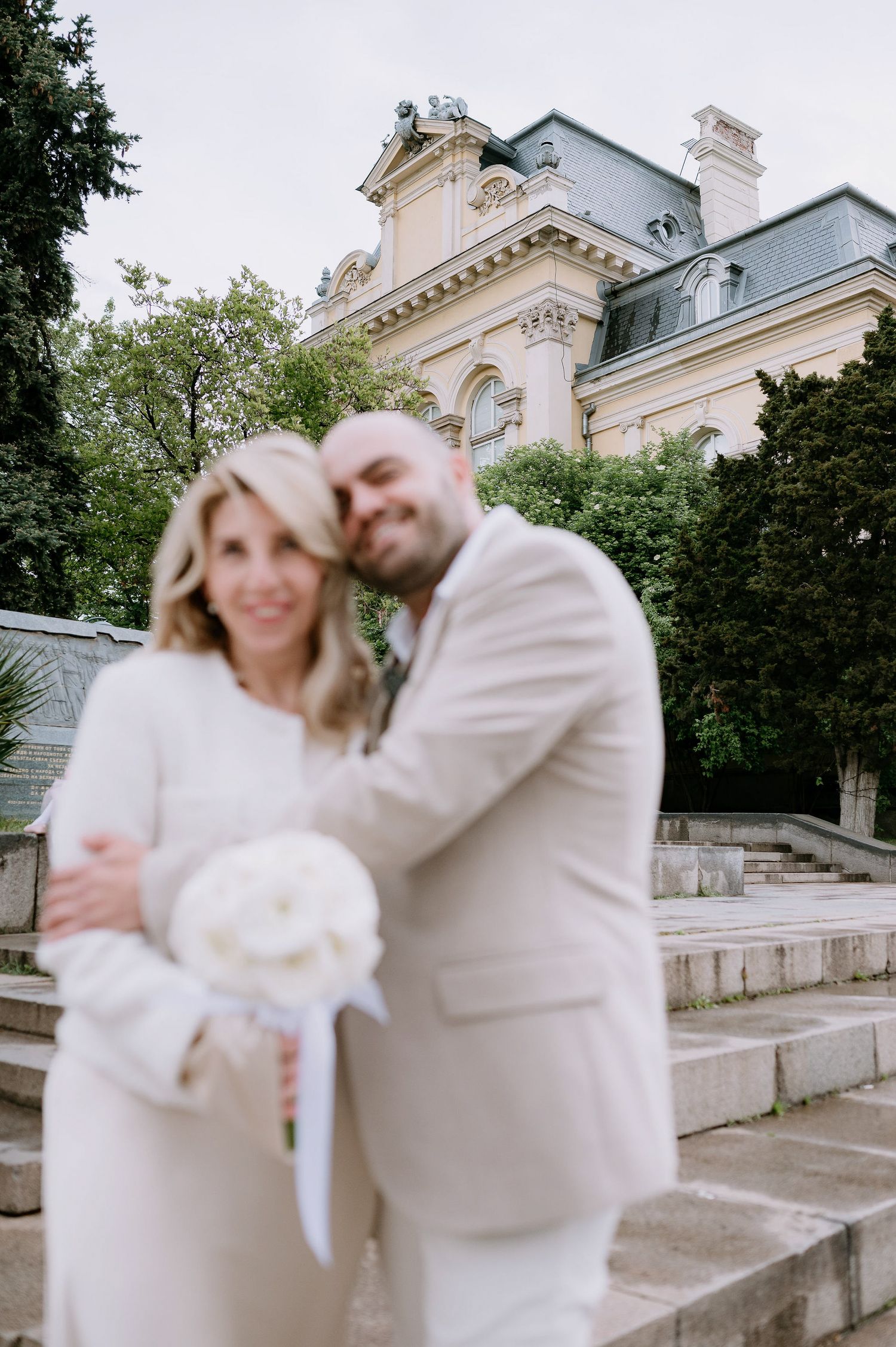 A couple in white clothing shares an intimate moment on stone steps near classical architecture.