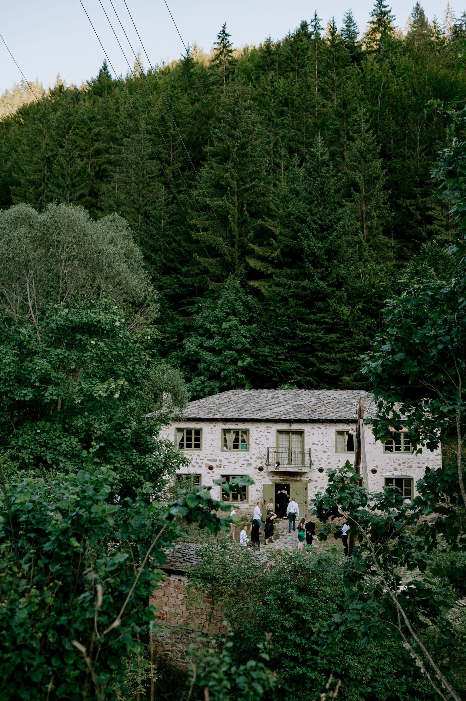 Stone house nestled in dense evergreen forest on mountainside with power lines overhead.
