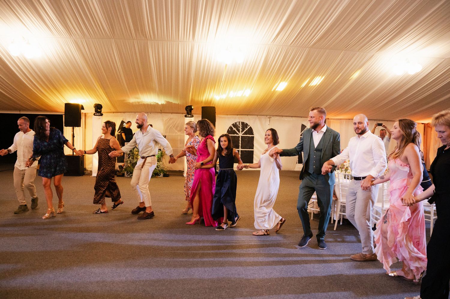 Wedding guests dance together under a white tent with warm lighting at an evening reception celebration.