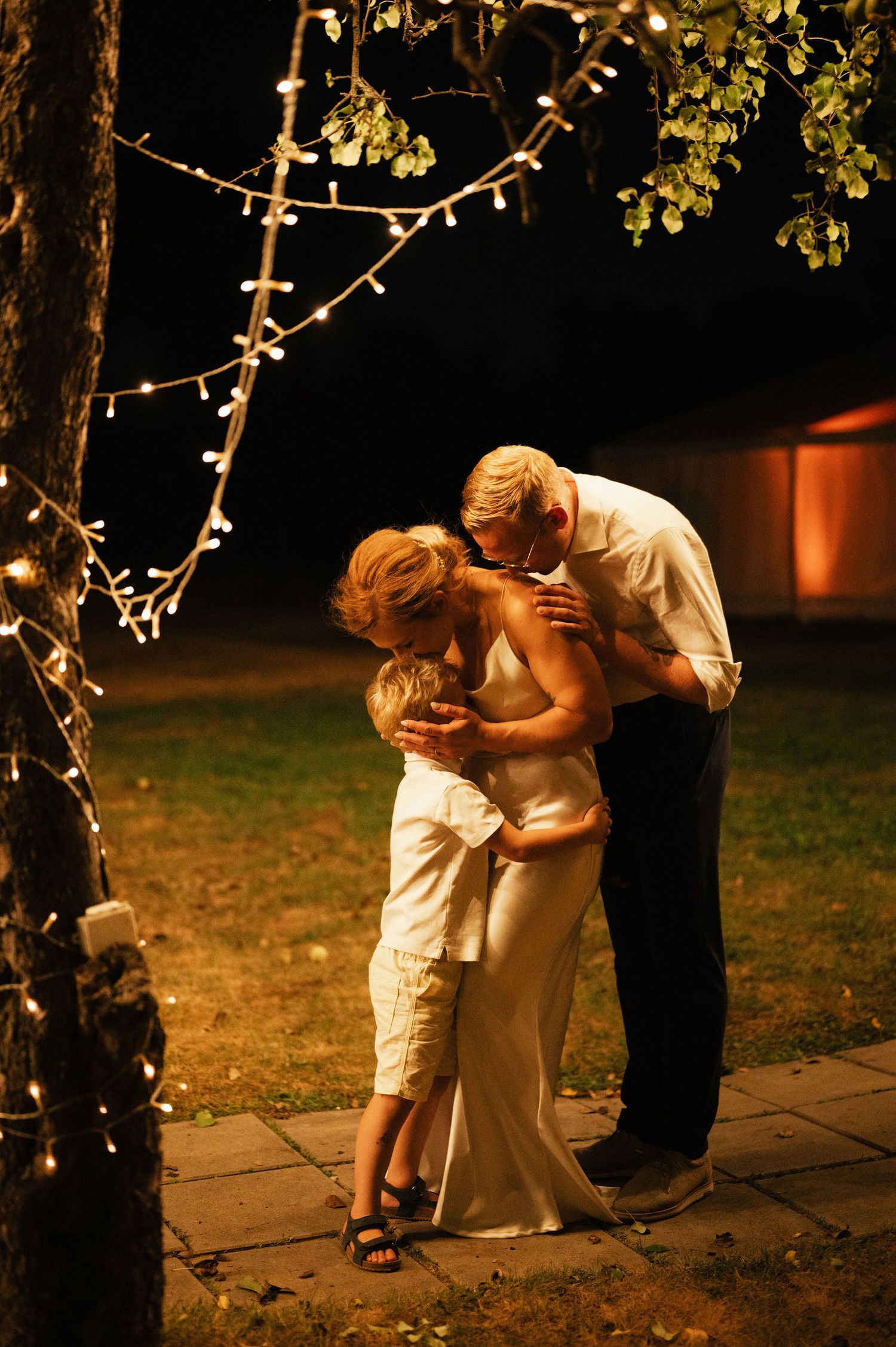 A romantic couple shares an intimate kiss at night under twinkling lights in a garden setting.