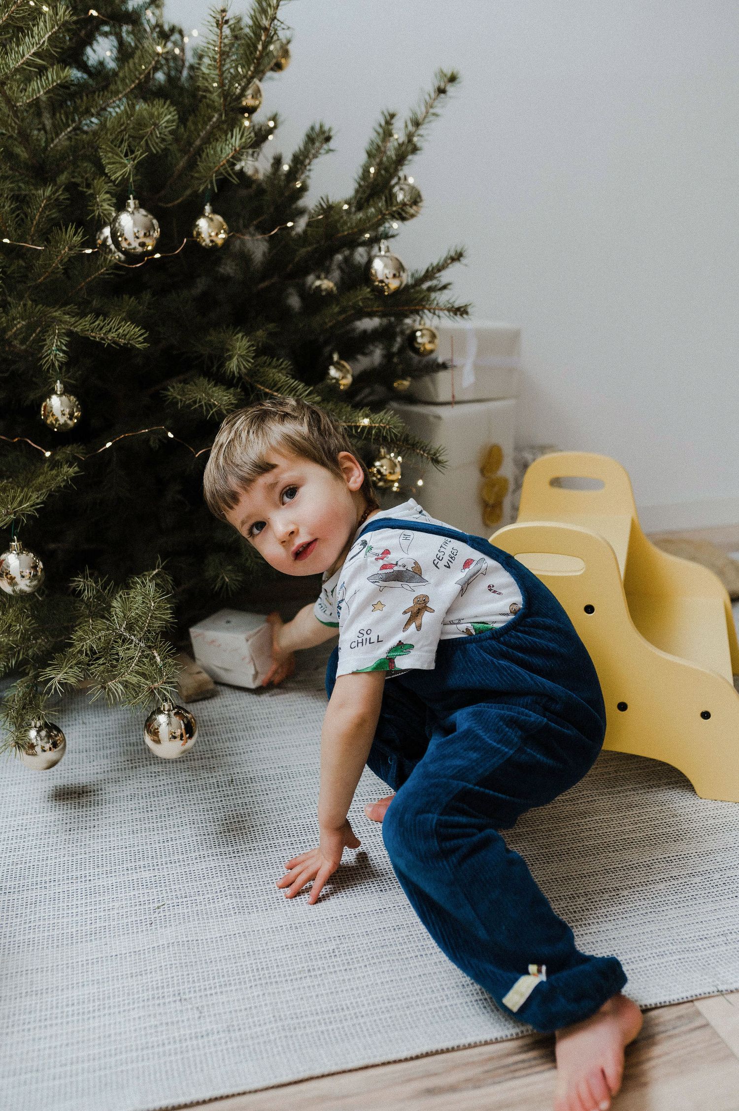 A child lays on a white carpet while playing near a Christmas tree decorated with silver ornaments.