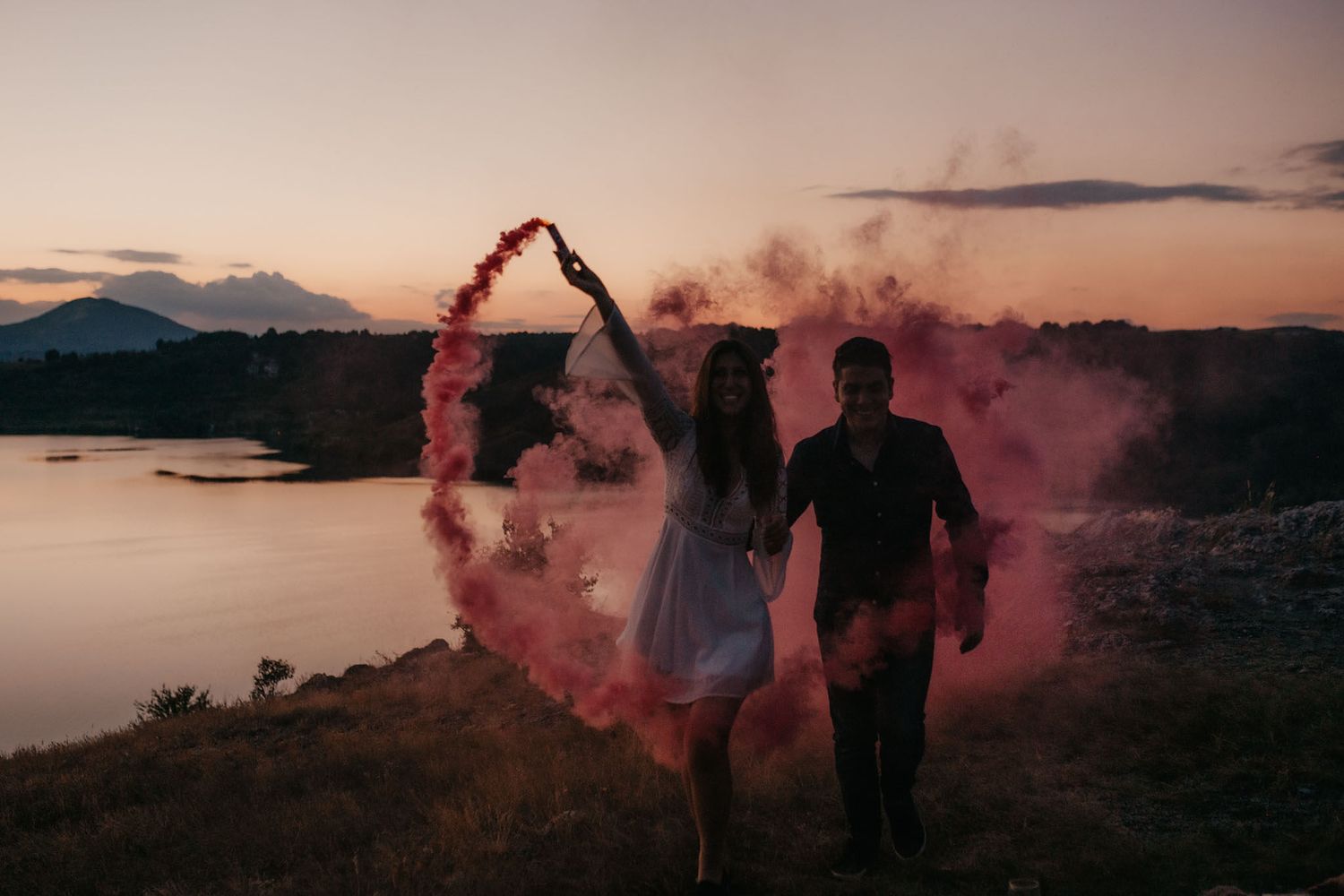 Silhouettes emerge through red smoke against a sunset lake backdrop.