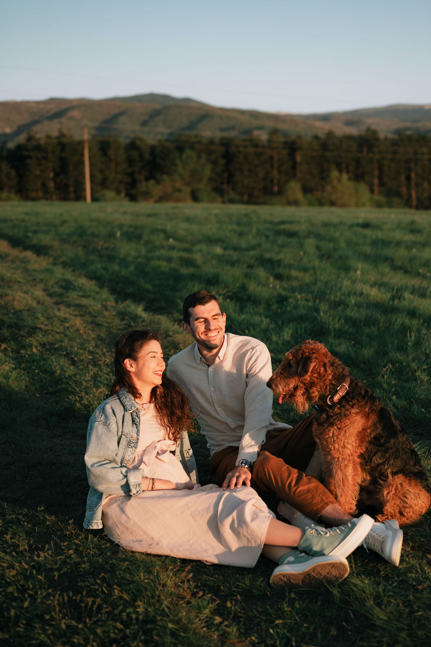 A couple relaxes together in a grassy field at sunset with their brown poodle dog.