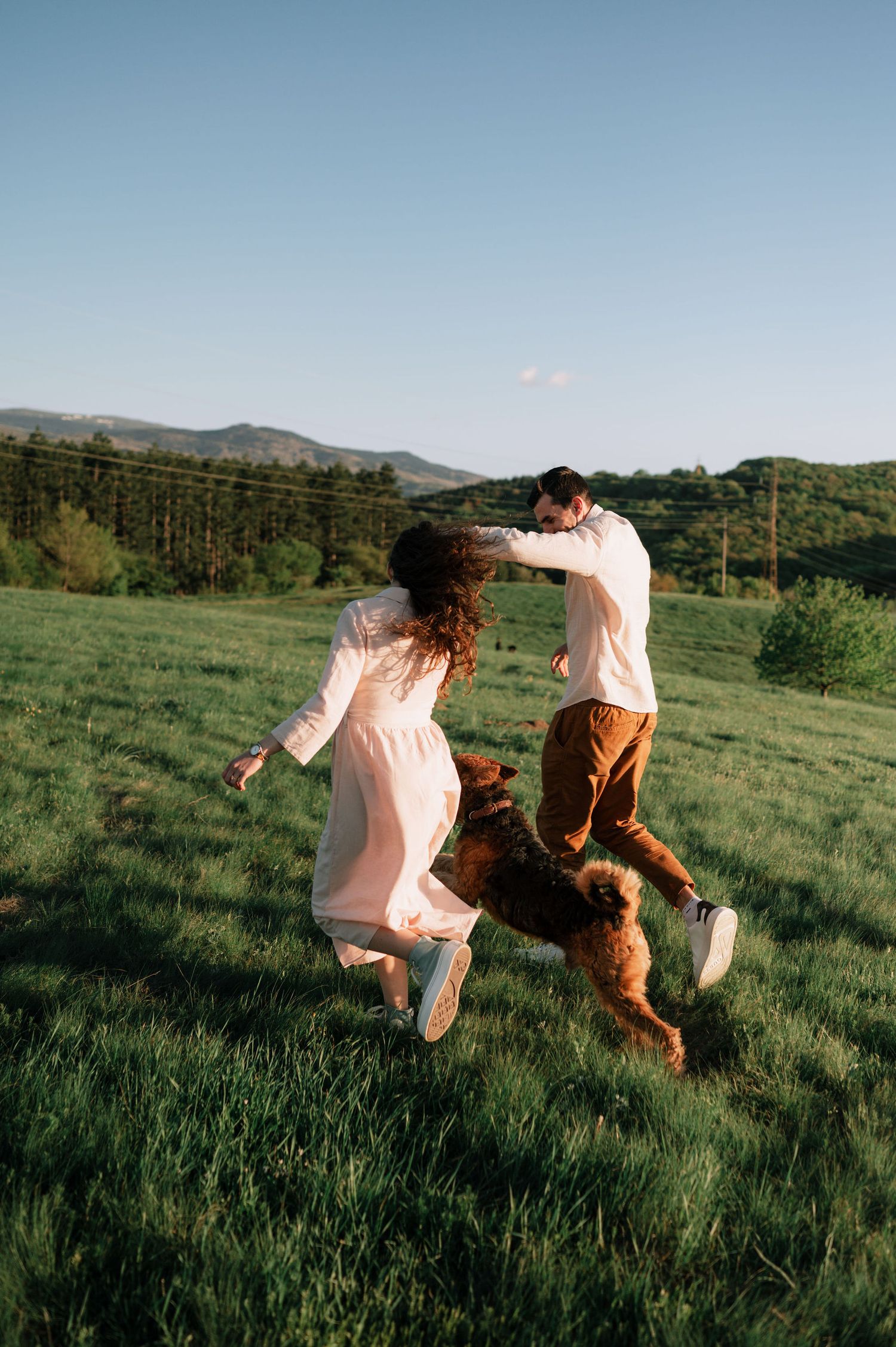 People in white dresses dance playfully with dog in green mountain field.