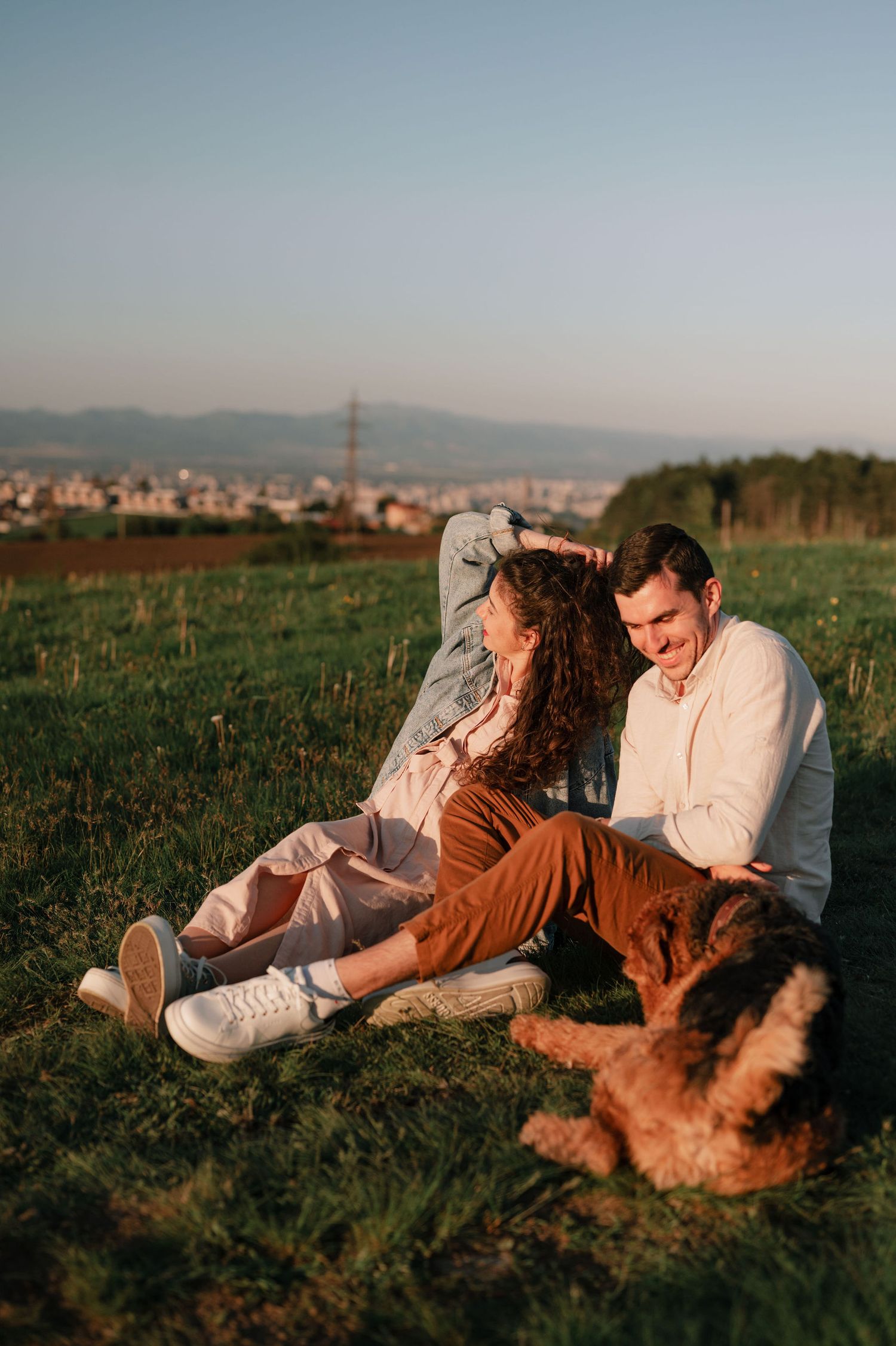 A couple relaxes in a grassy meadow at sunset with their golden retriever dog enjoying a peaceful moment together.