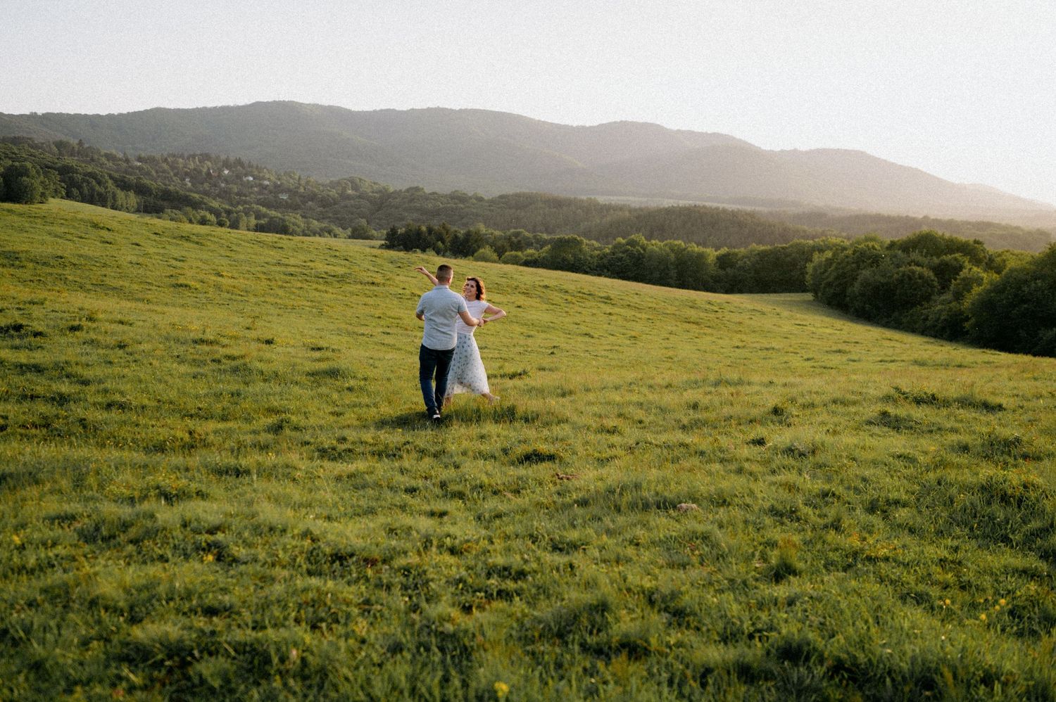 A couple walks hand in hand through a green mountain meadow at sunset.