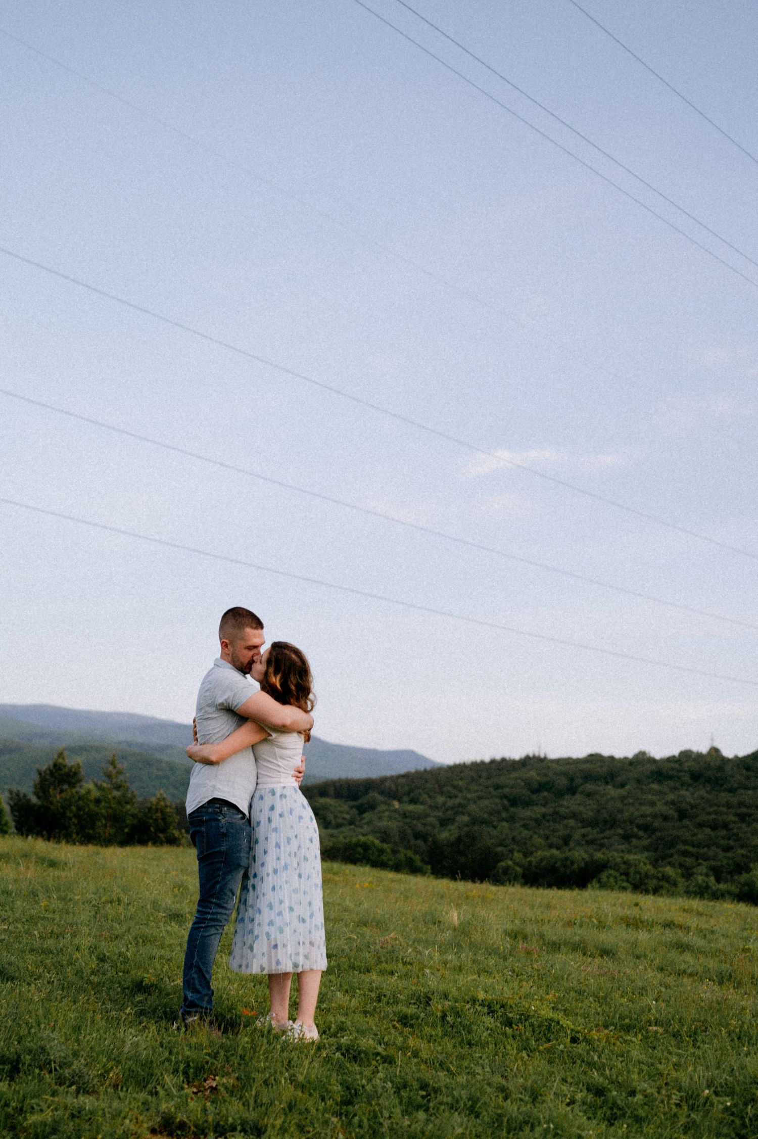 A romantic couple embraces in a scenic mountain meadow filled with wildflowers and rolling hills.