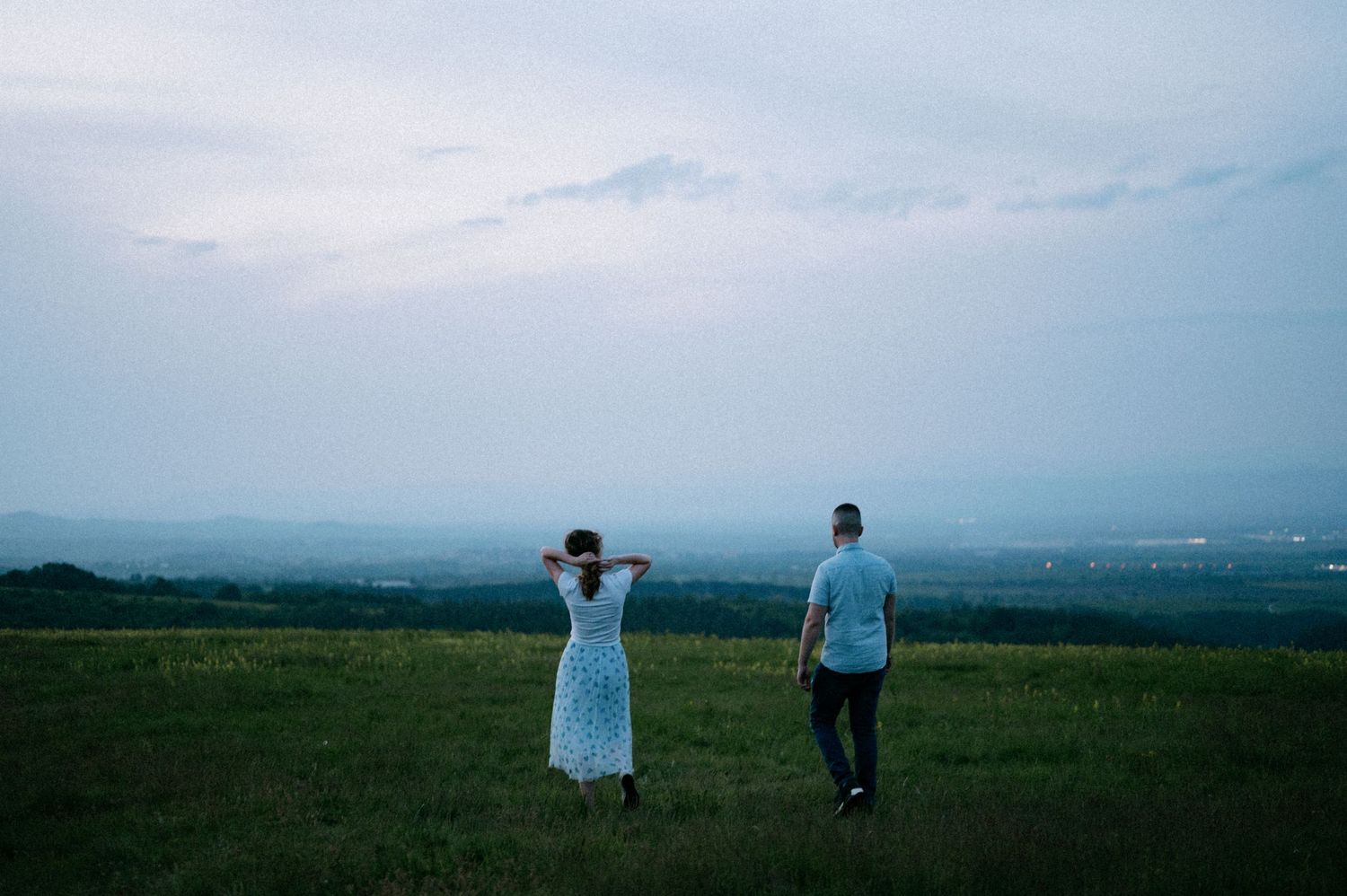 Two people stand on a hilltop overlooking a vast landscape at dusk.
