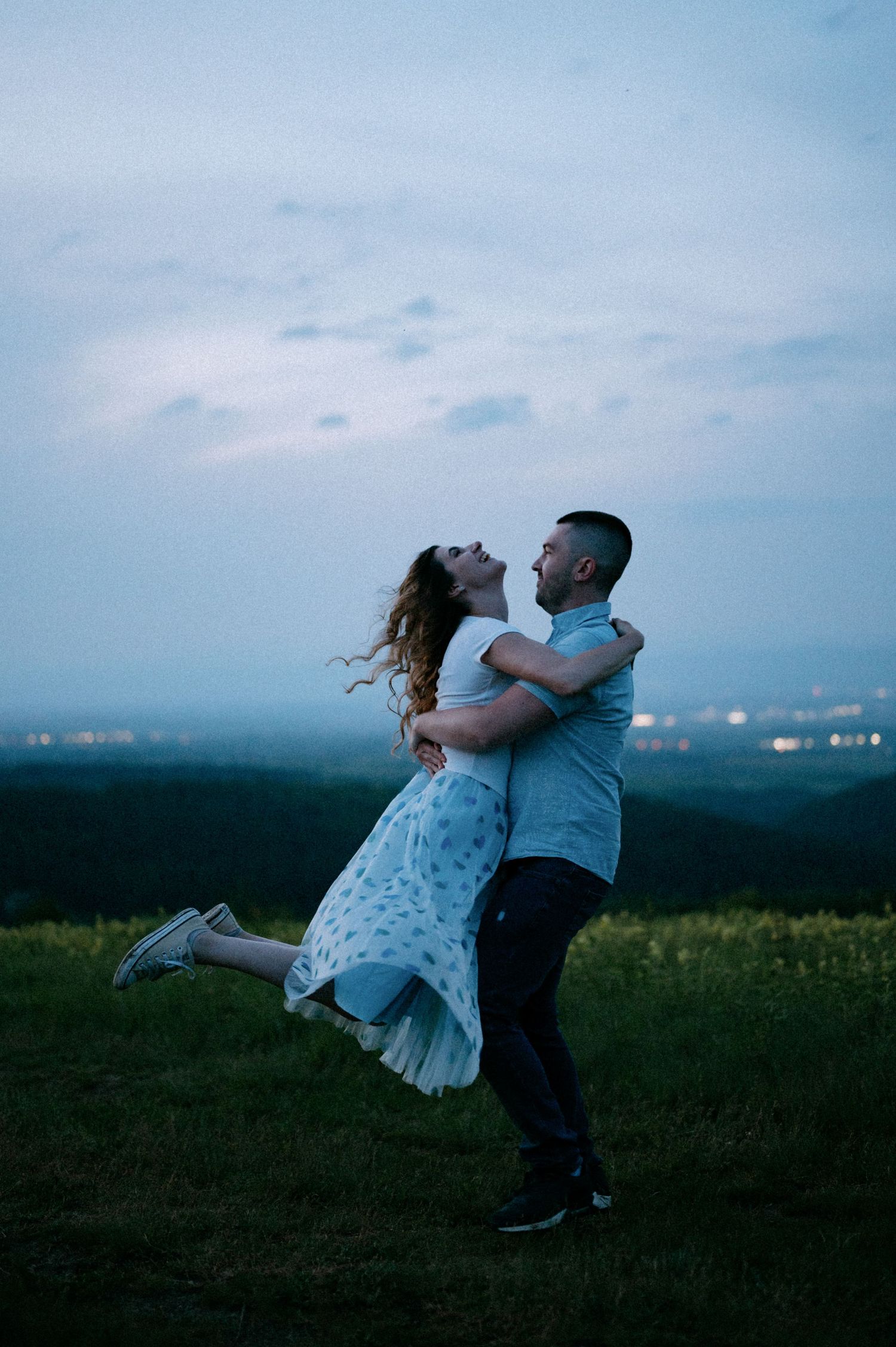 A couple dances and spins together on a hilltop at dusk with a city skyline visible in the distance.