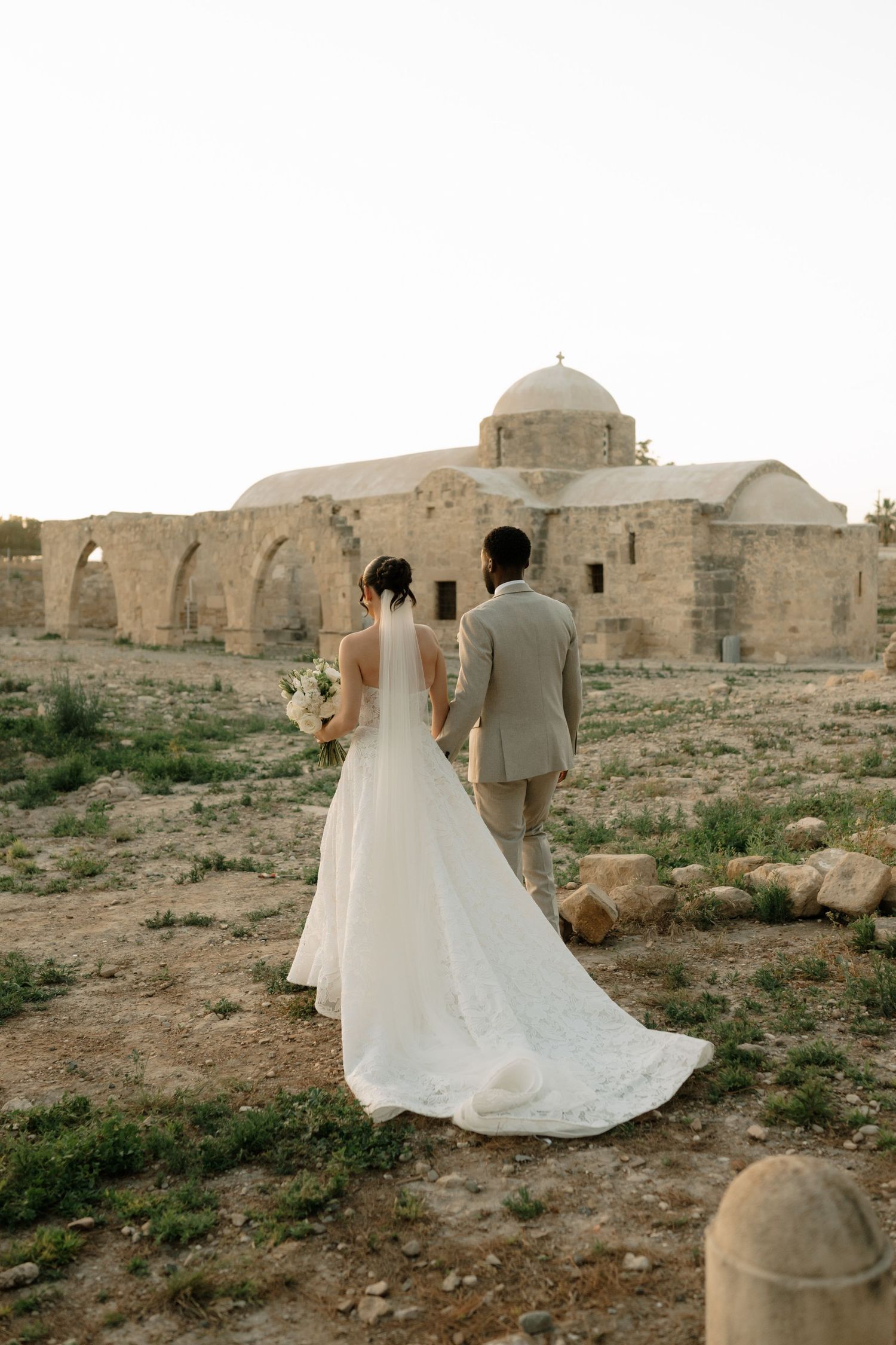 Bride and groom walking toward ancient stone church ruins during golden hour wedding photoshoot.