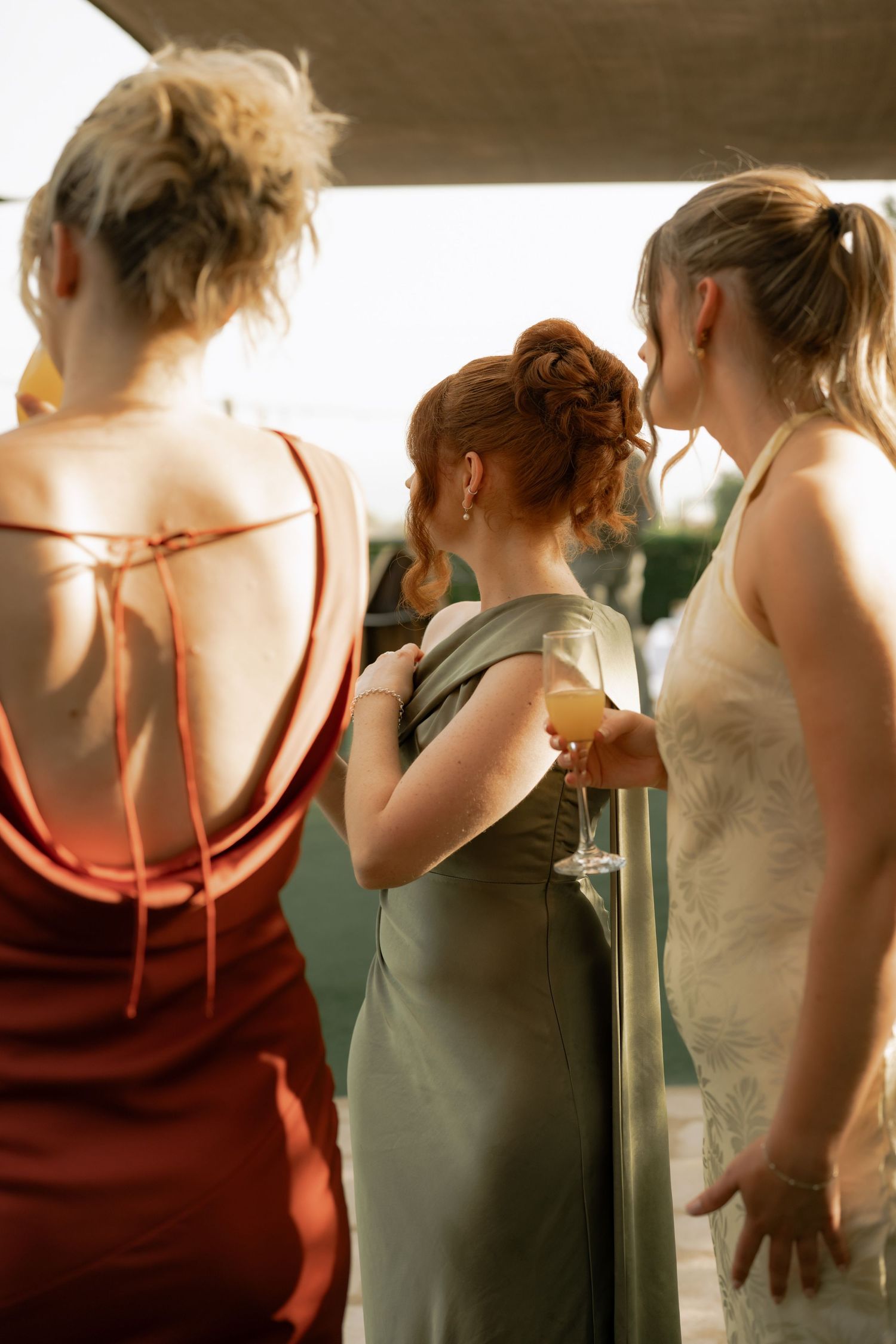 Three women in elegant dresses standing on a balcony, one holding a champagne glass at a formal event.