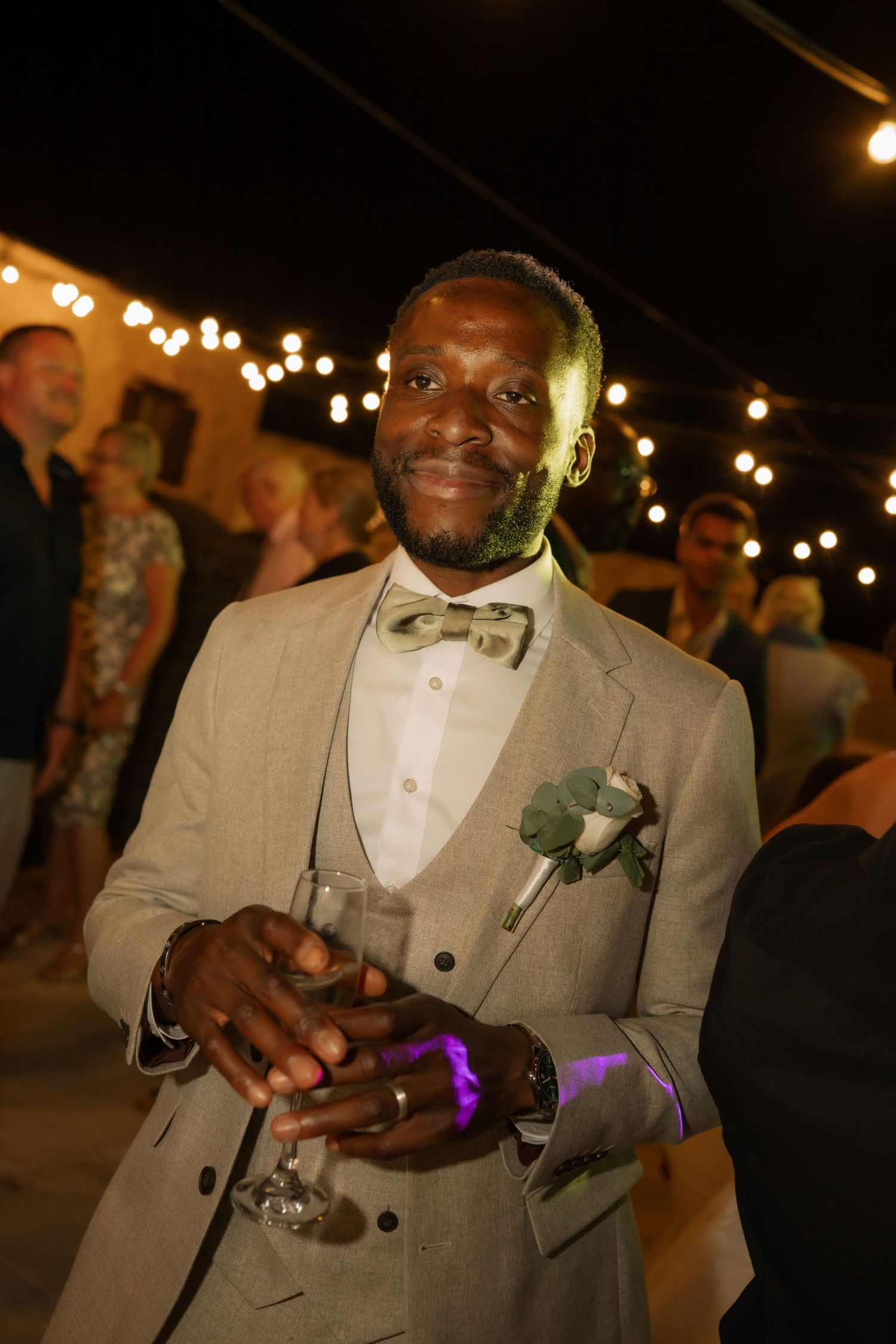 A well-dressed man in a tan suit and bow tie holds a wine glass at an elegant outdoor evening wedding reception.