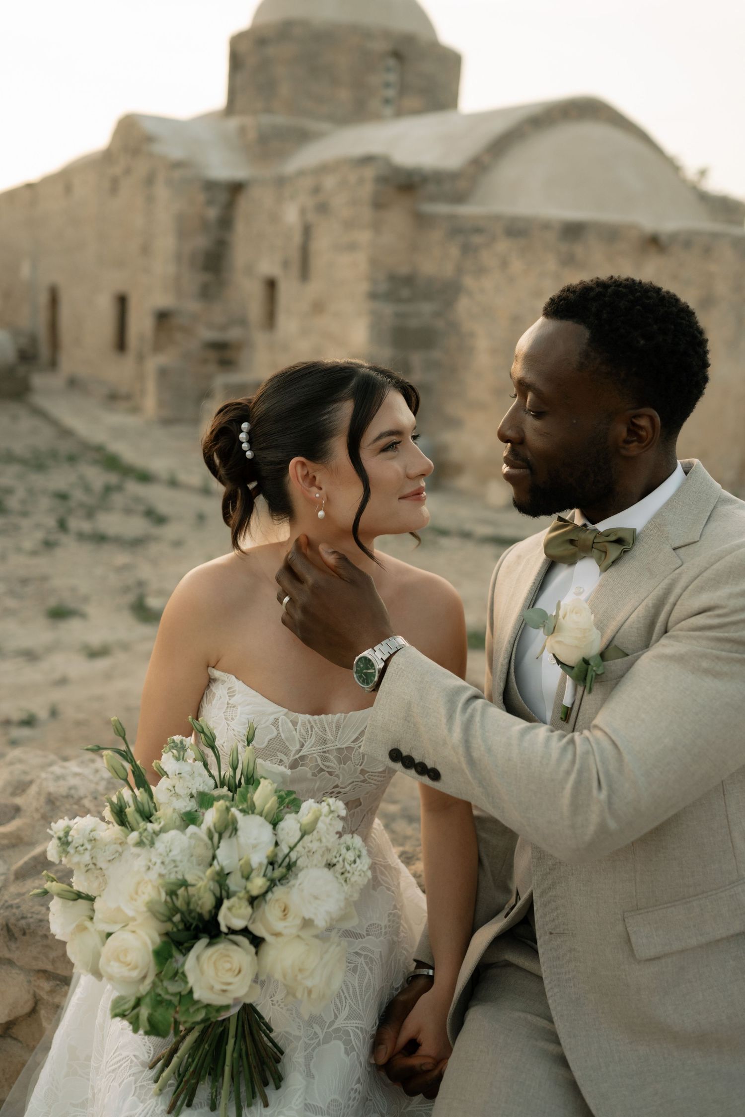 Interracial couple in wedding attire embrace near ancient stone ruins, bride holding white floral bouquet at sunset.