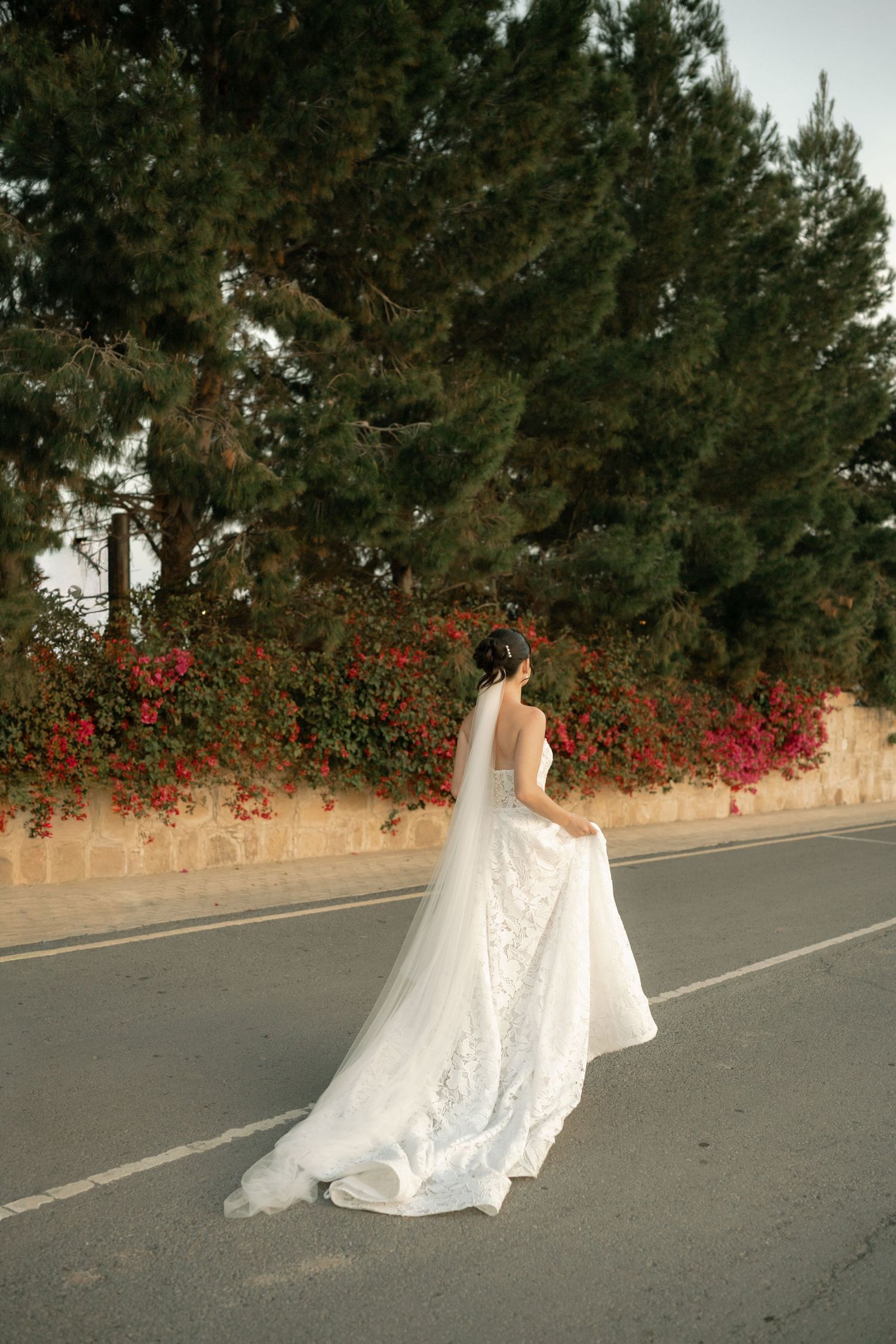 Bride in elegant white lace wedding gown with long veil stands on road surrounded by lush green trees and red flowers.
