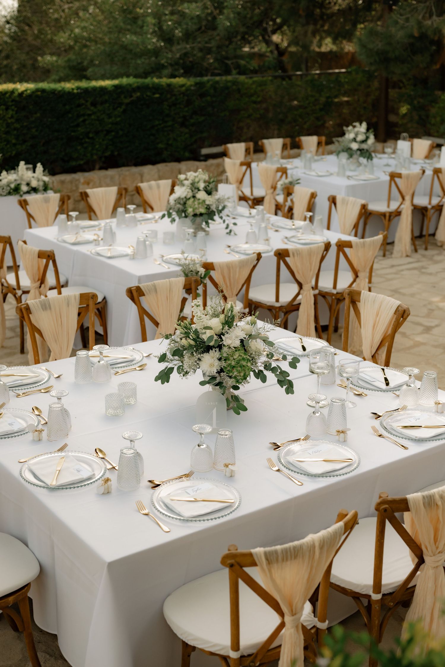 Elegant outdoor wedding reception with white tablecloths, wooden cross-back chairs, and floral centerpieces in a garden setting.