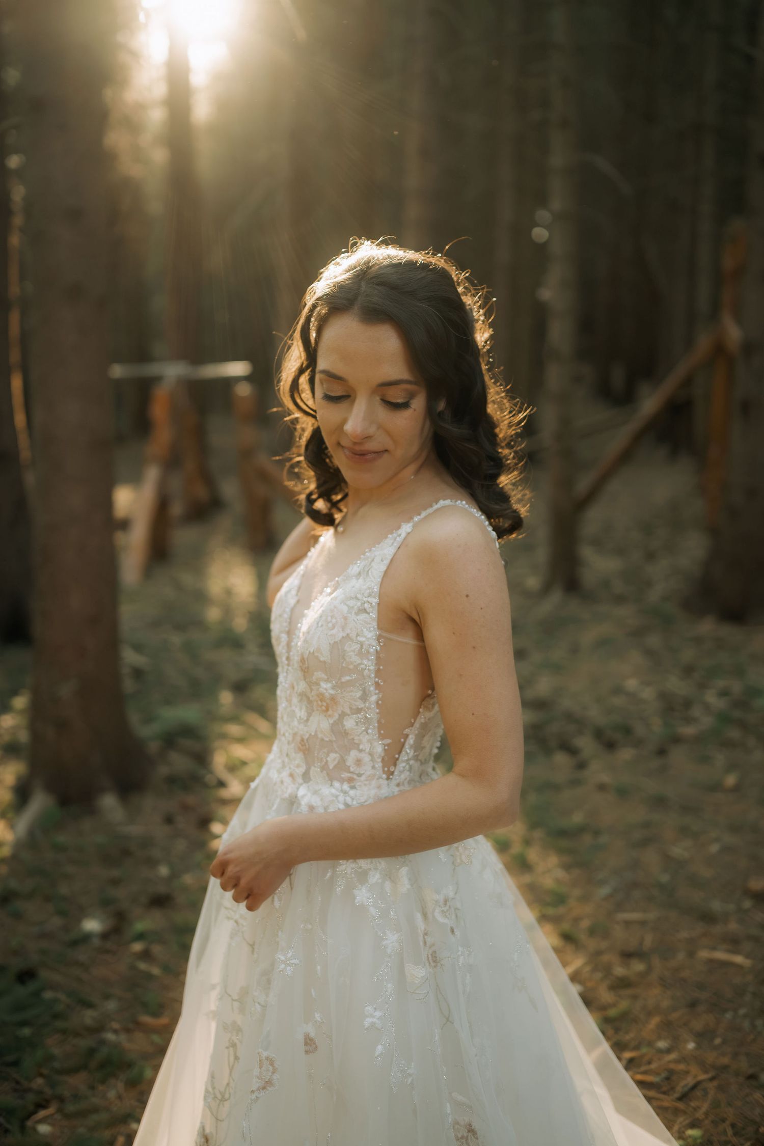 Bride in lace wedding gown standing in sunlit forest, looking down gracefully with golden backlight.