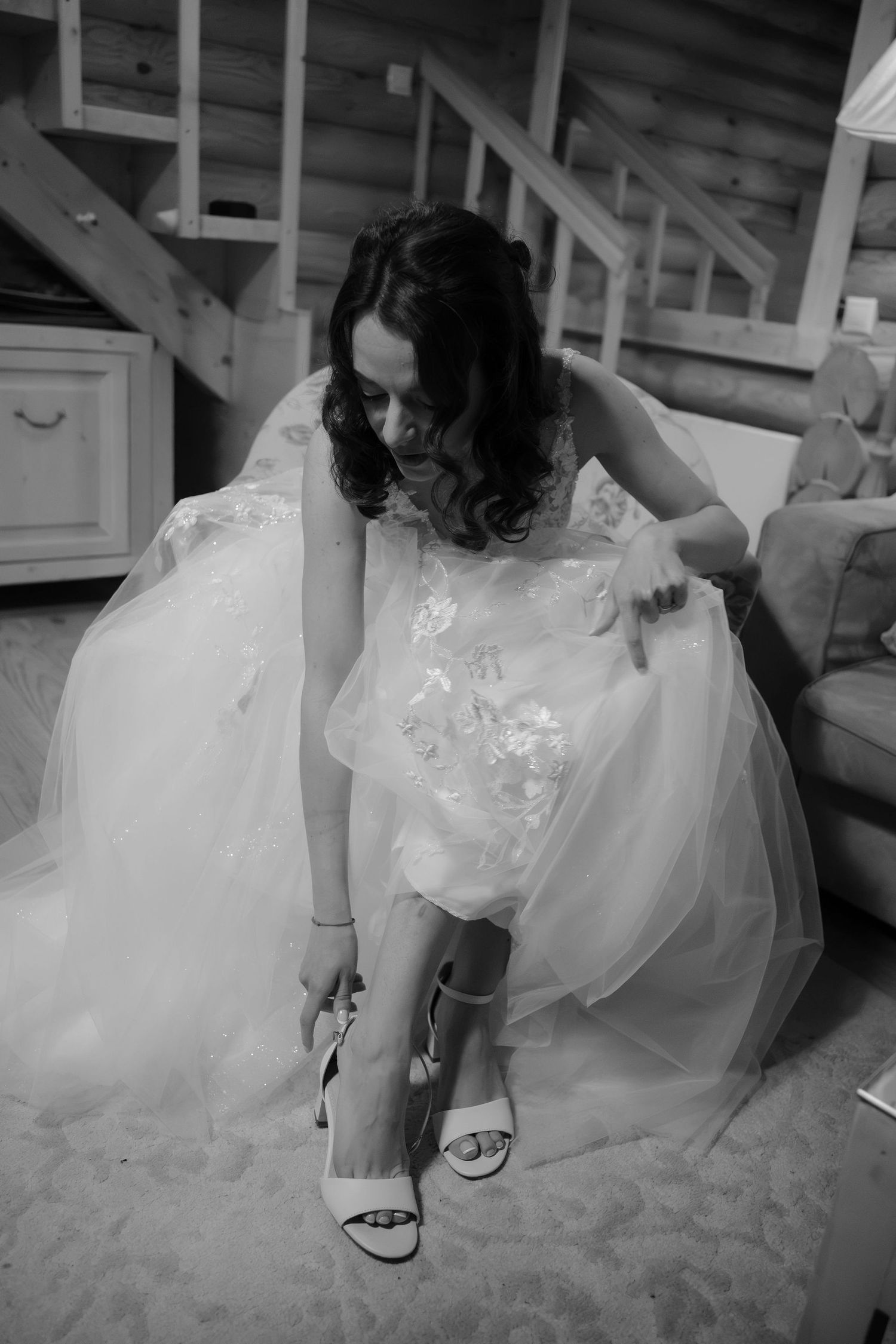 Bride in white wedding gown adjusting her strappy heels in black and white photo inside a rustic home.