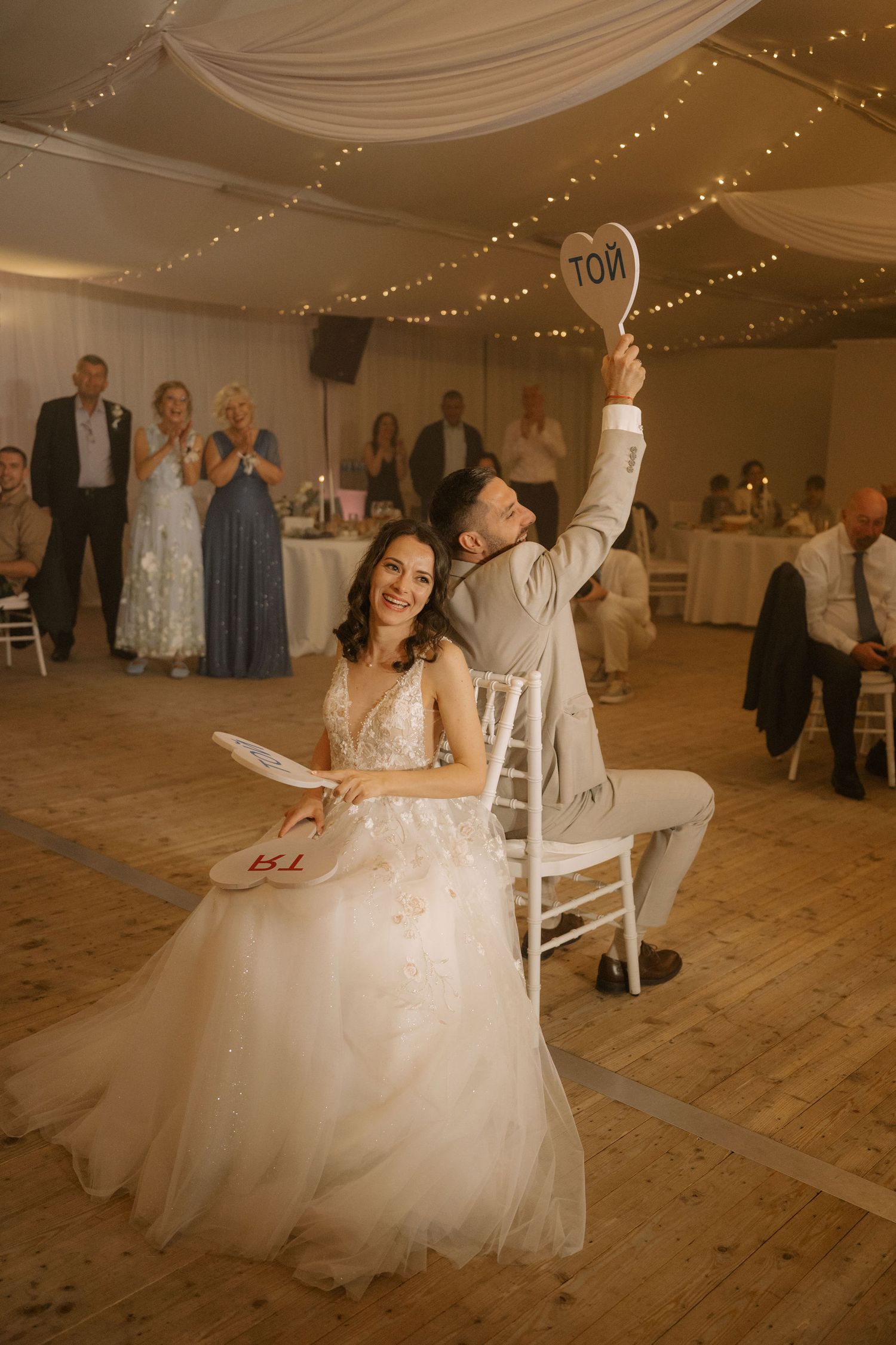 Bride and groom holding heart-shaped paddles during a fun wedding game in a decorated reception venue with fairy lights.