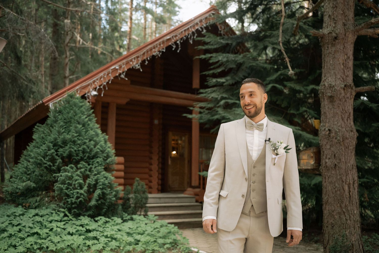 Groom in beige suit with boutonniere stands outdoors near rustic log cabin surrounded by pine trees.