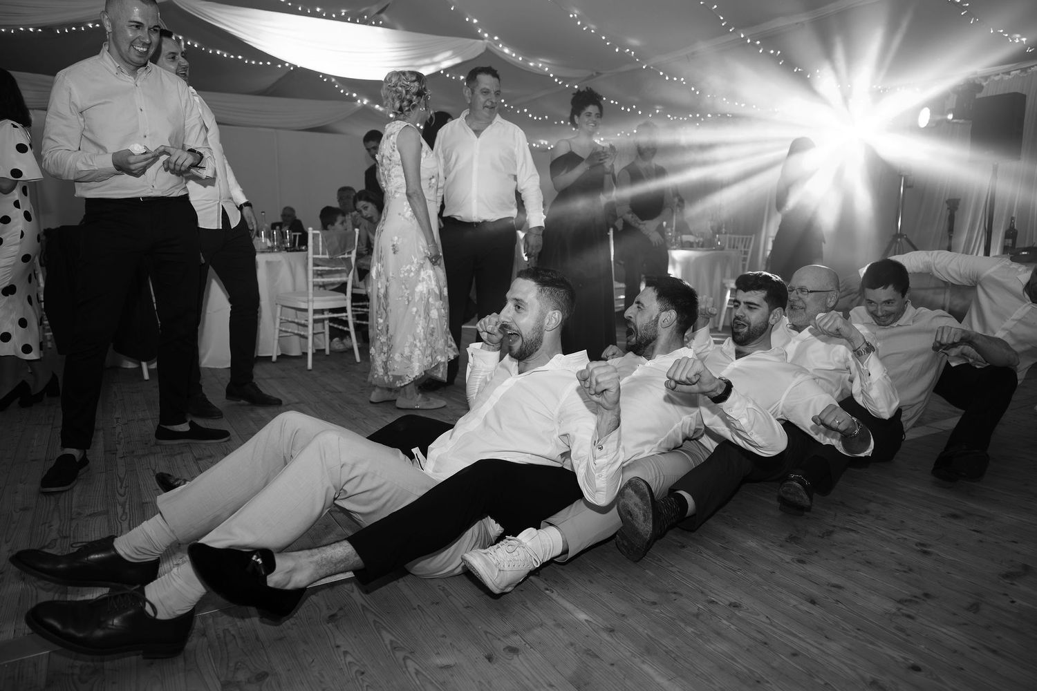 Black and white photo of groomsmen performing a fun train dance move on the wedding reception dance floor.