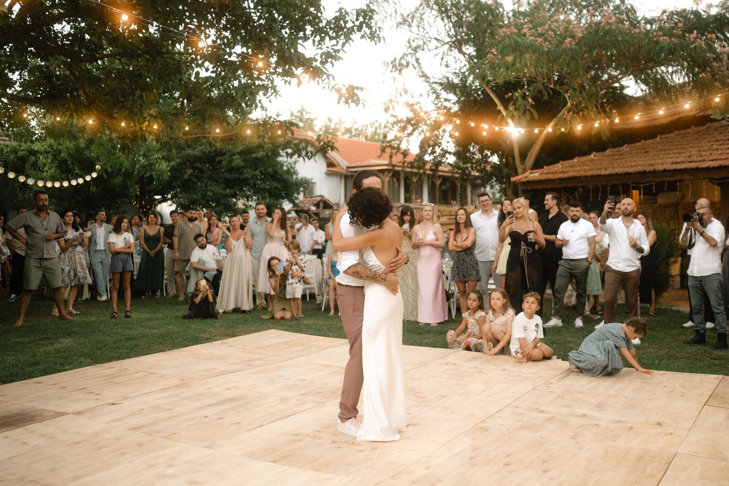 Couple shares first dance at outdoor wedding reception with string lights and guests watching on a wooden dance floor.