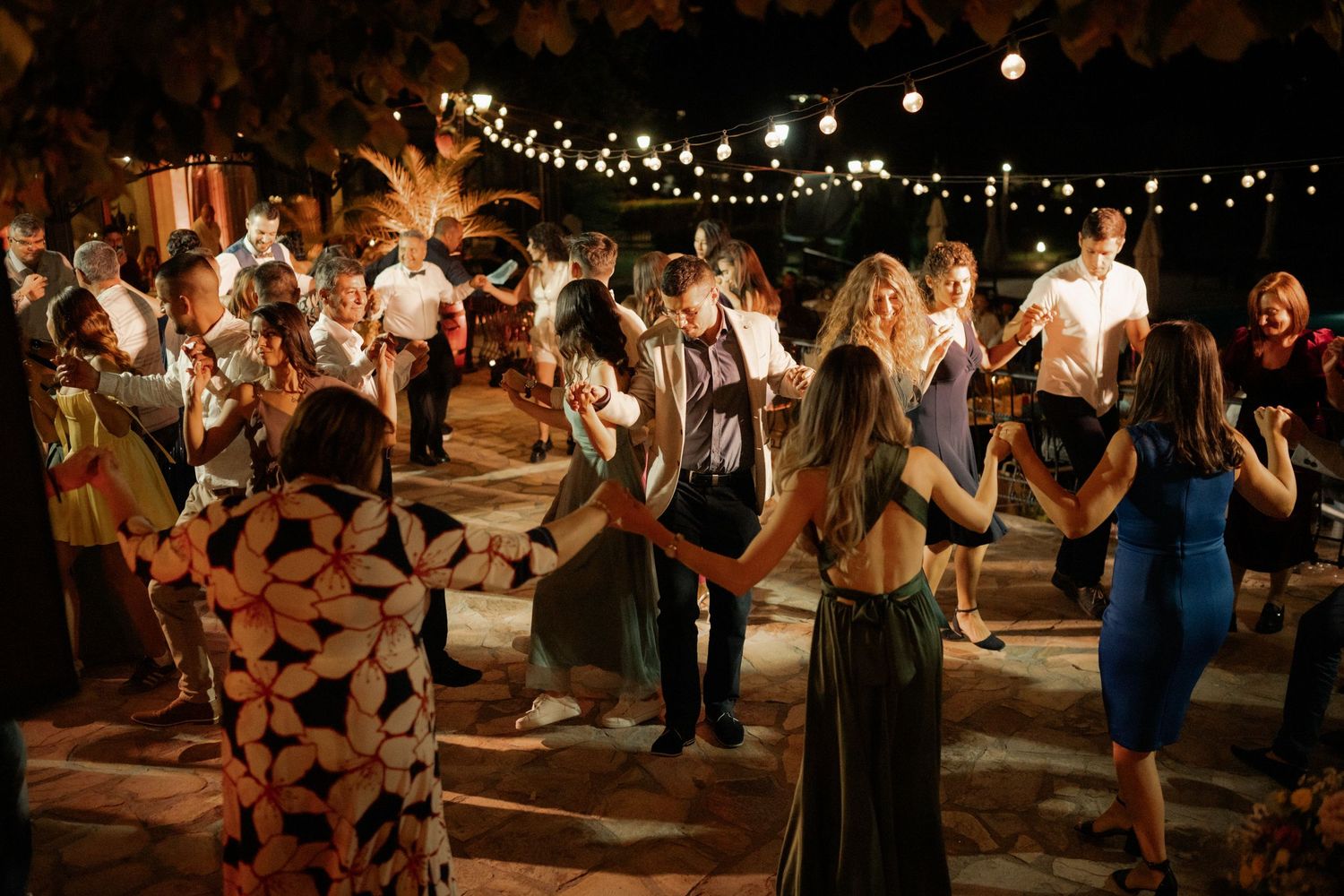 People dancing and celebrating at an outdoor evening reception under string lights and trees.