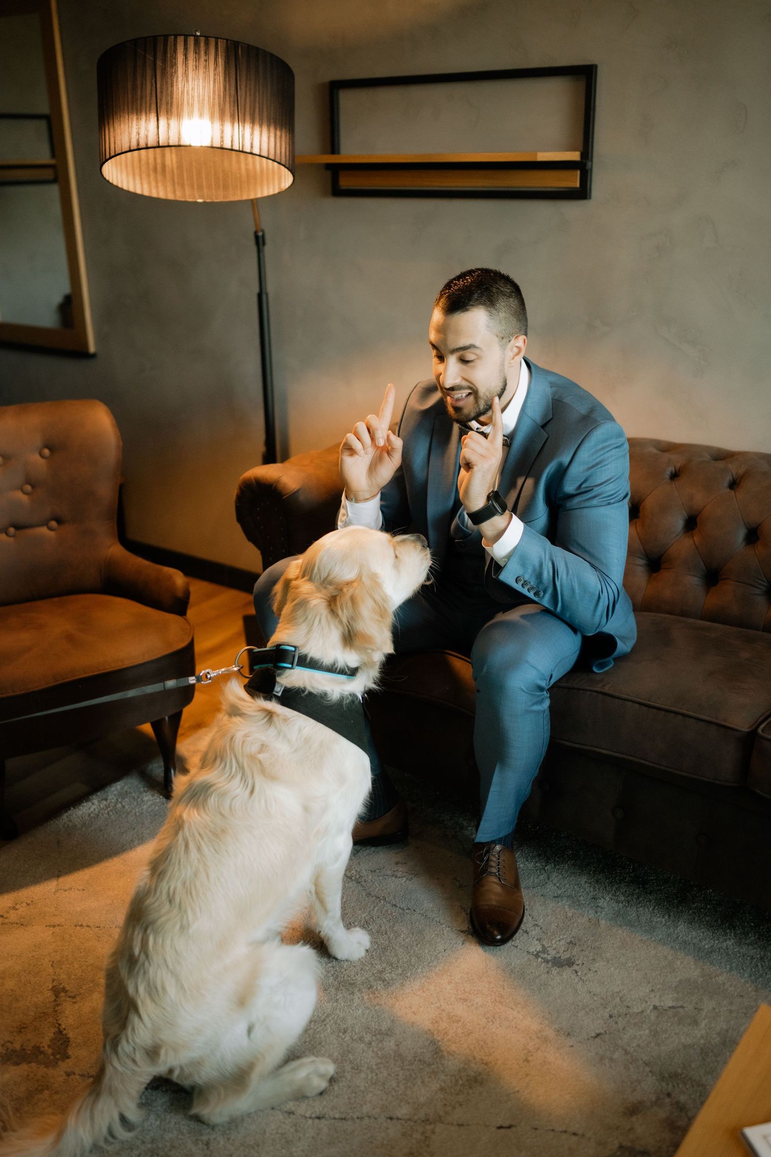 A person in a blue suit sits on a brown couch sharing a tender moment with a fluffy golden retriever dog.