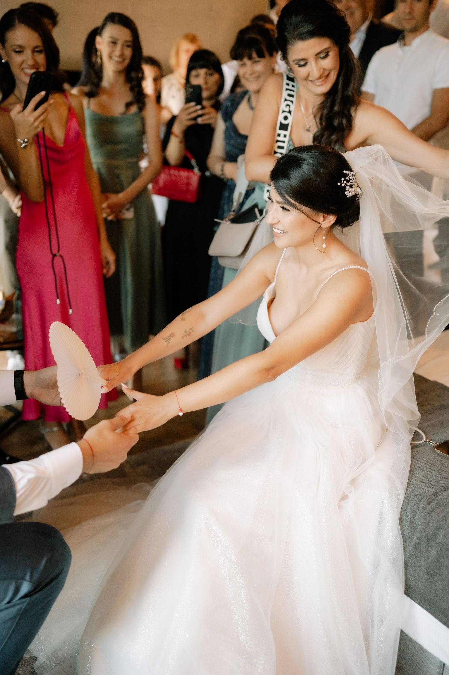 A bride in a flowing white wedding dress and veil dances joyfully at a reception surrounded by guests.