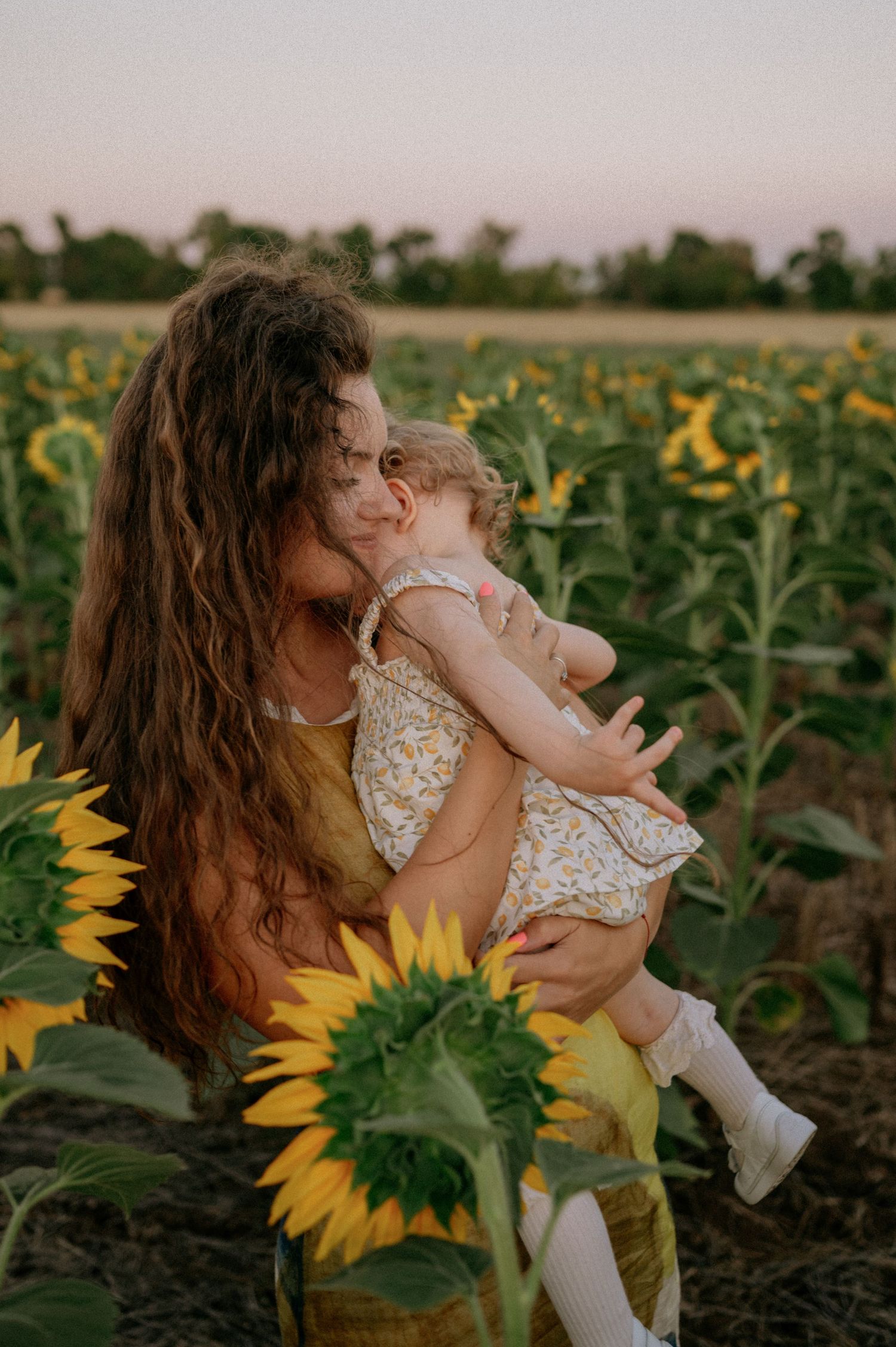 A mother with curly hair holds her toddler in a sunflower field at golden hour.