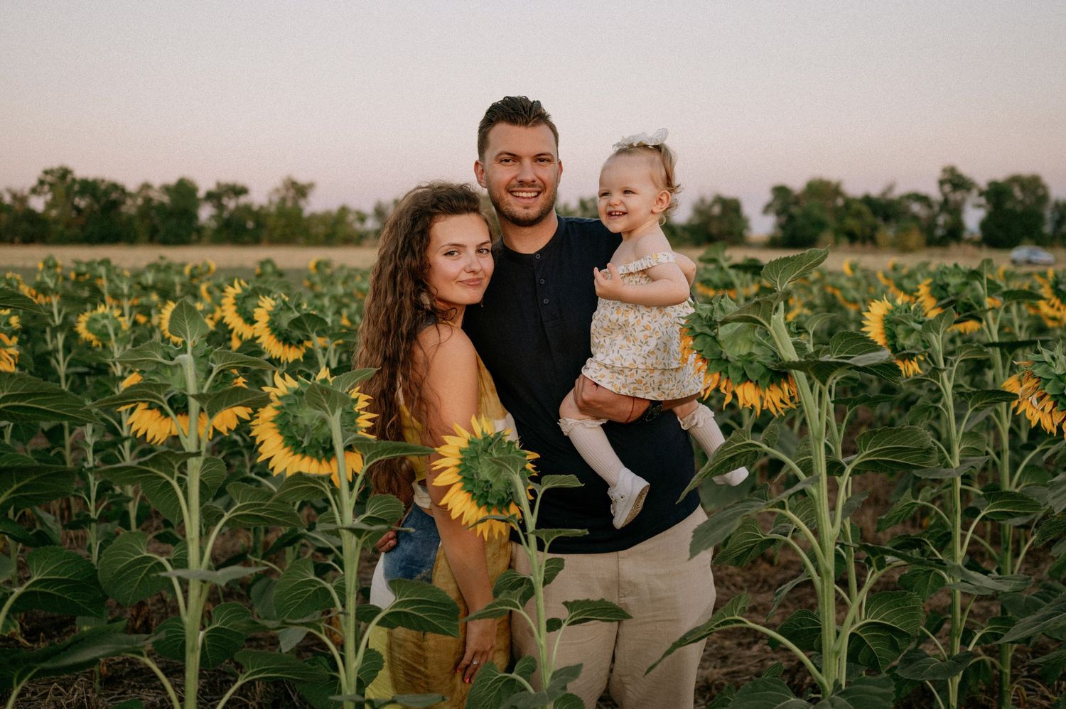 Family of three smiling in a sunflower field at sunset, parents holding toddler among tall yellow blooms.