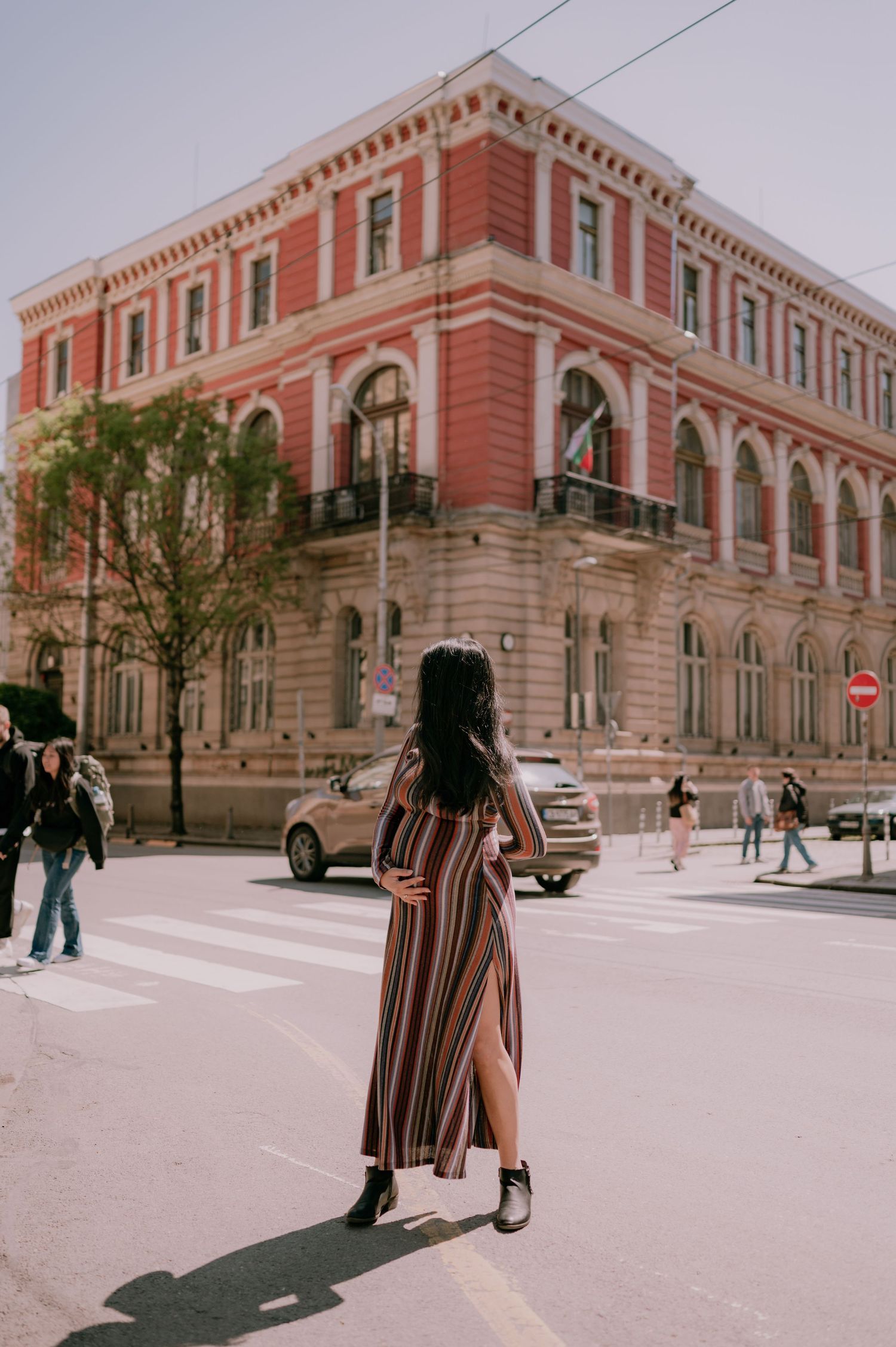 Woman in striped maxi dress crossing street near historic red building on sunny day.