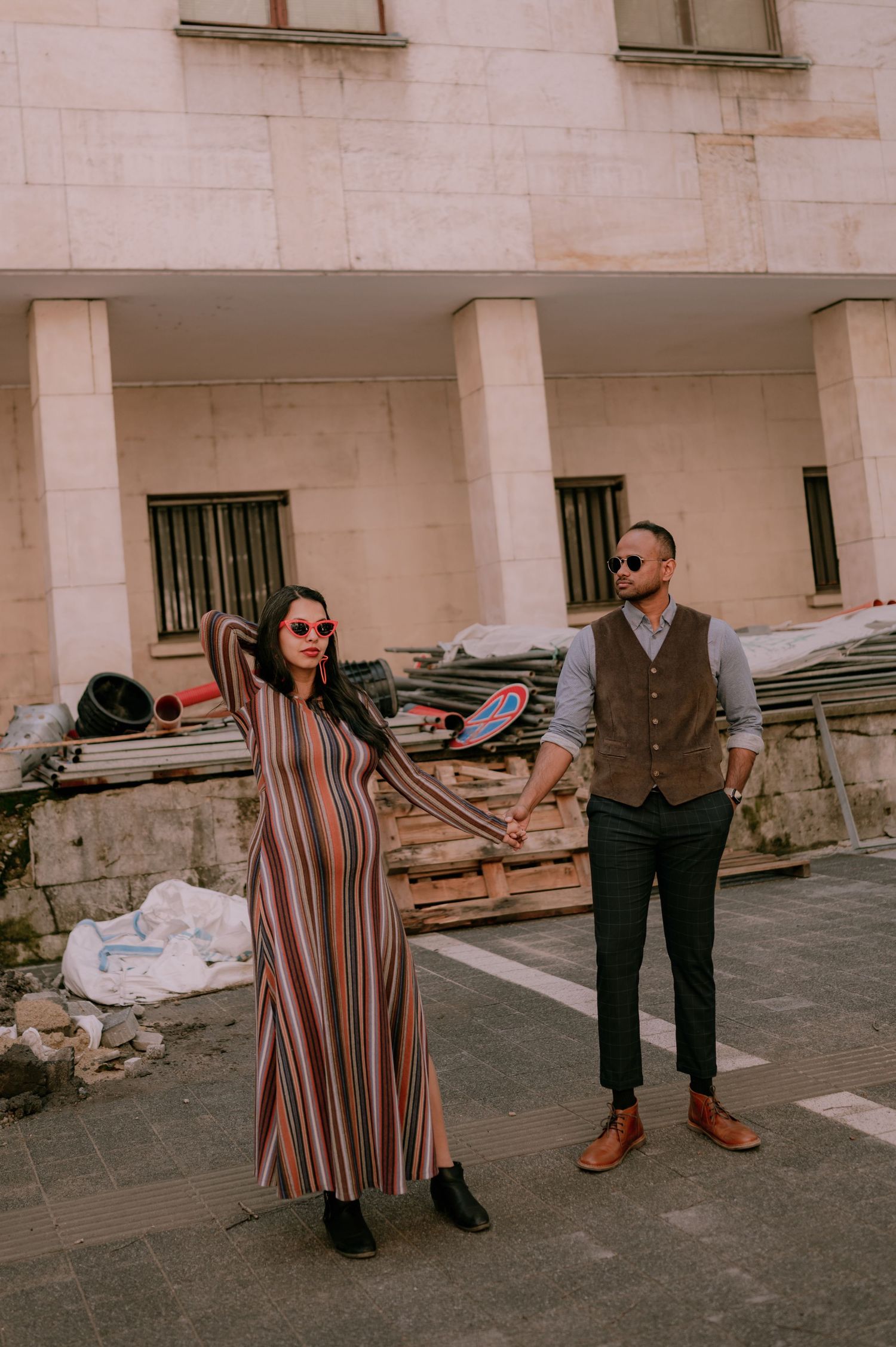 Stylish couple holding hands in front of a building under renovation, woman in striped dress, man in vest and trousers.
