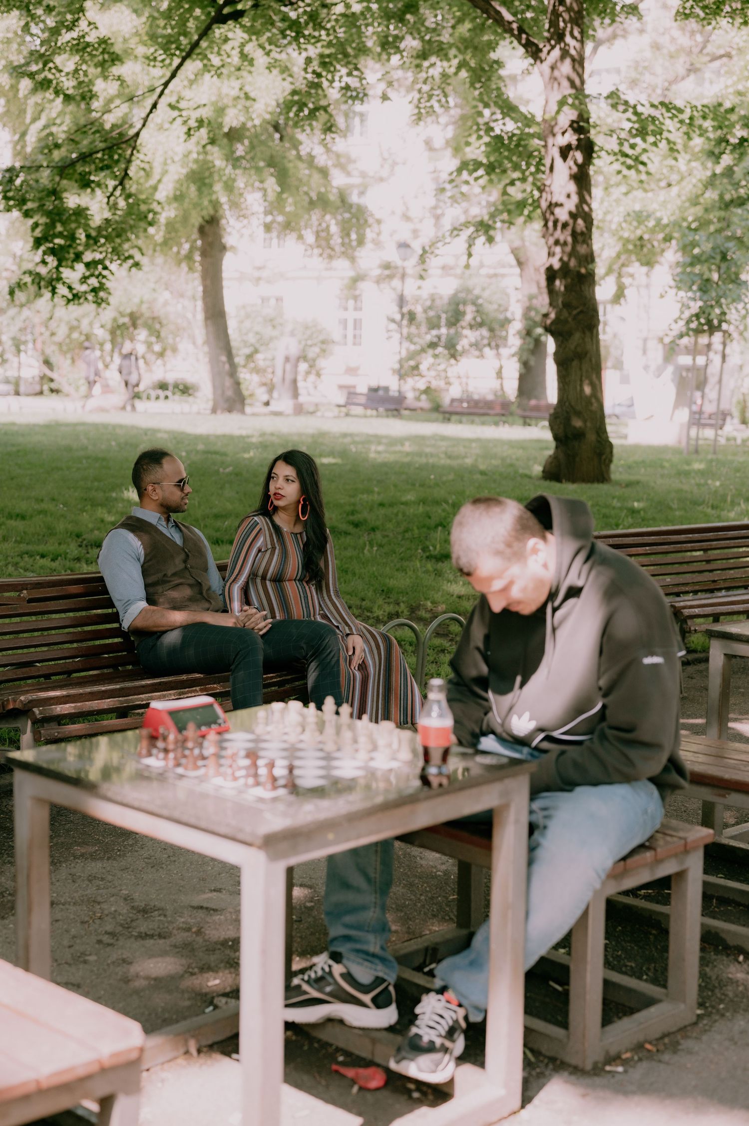 Two people sit on a park bench watching a man play chess at an outdoor table surrounded by lush green trees.
