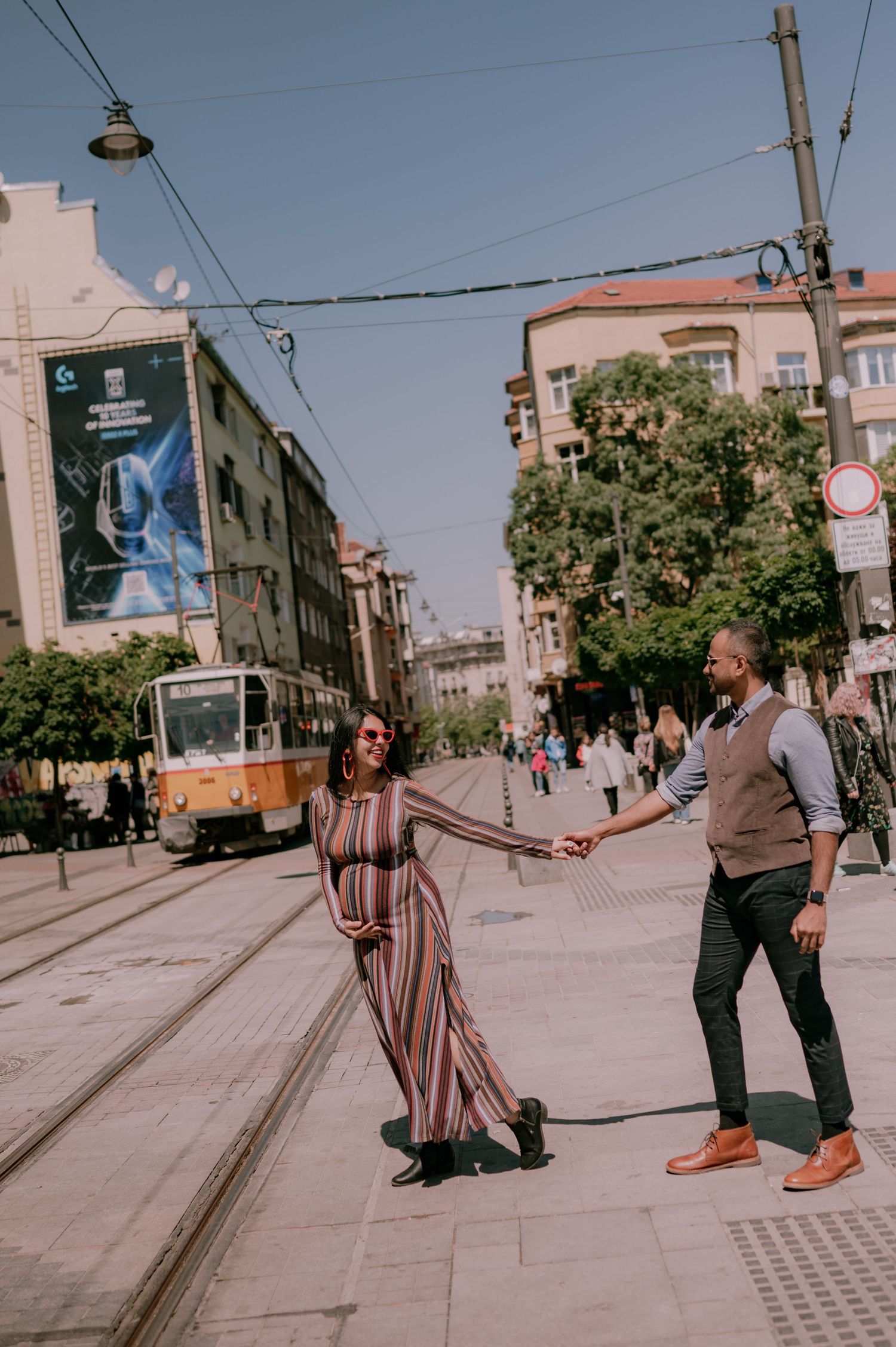 Pregnant woman in striped dress holds hands with man on urban street with tram tracks and yellow tram in background.