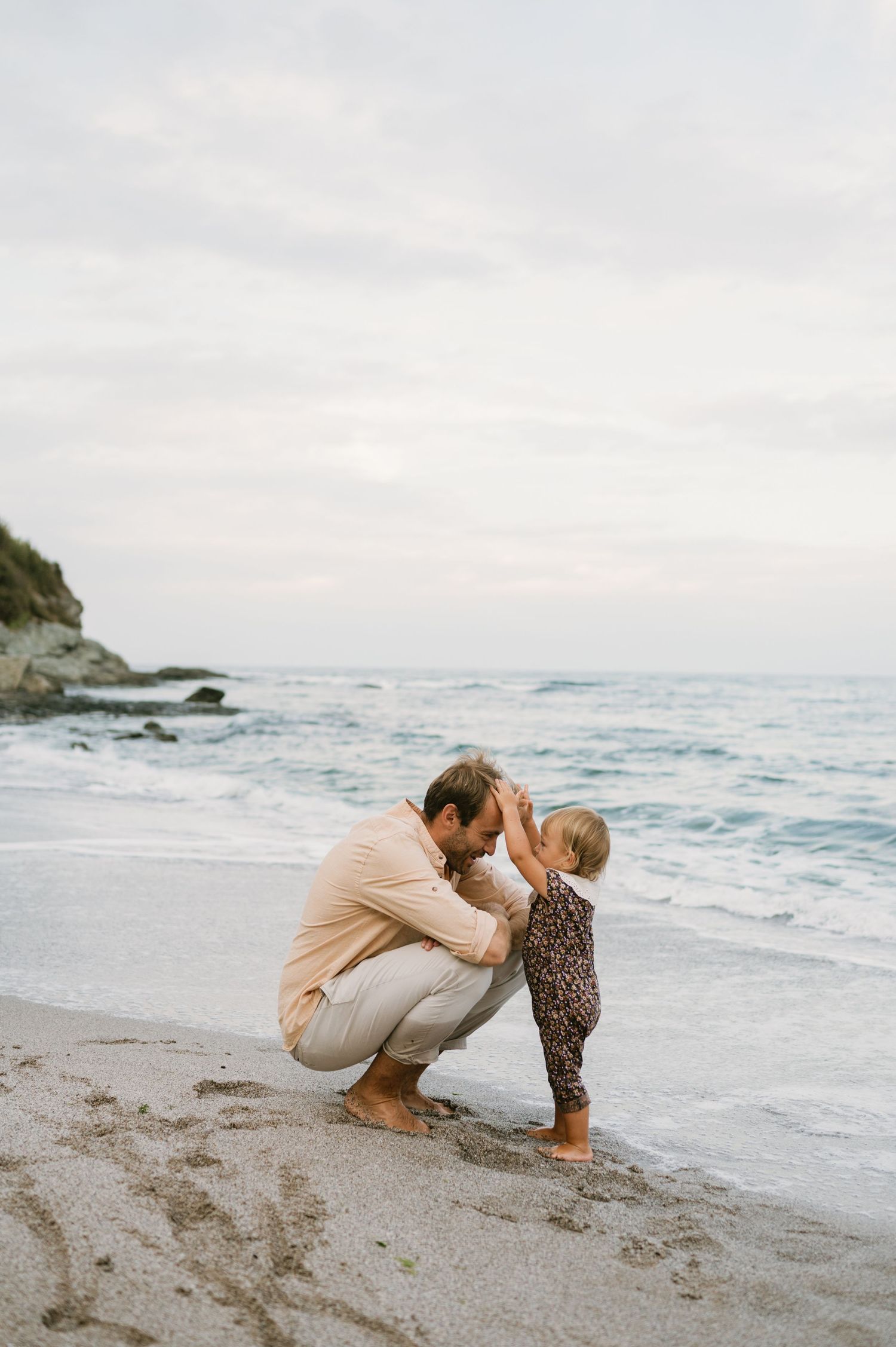 Father crouching on sandy beach tenderly interacting with toddler near ocean waves at dusk.