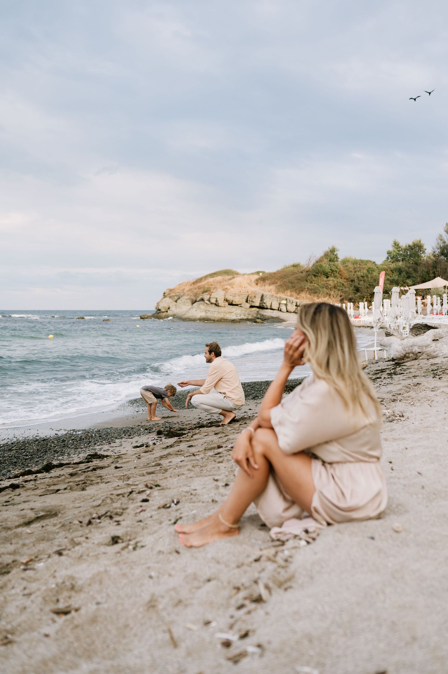 Couple relaxing on a sandy beach with rocky coastline, woman sitting in foreground, man crouching near waves.