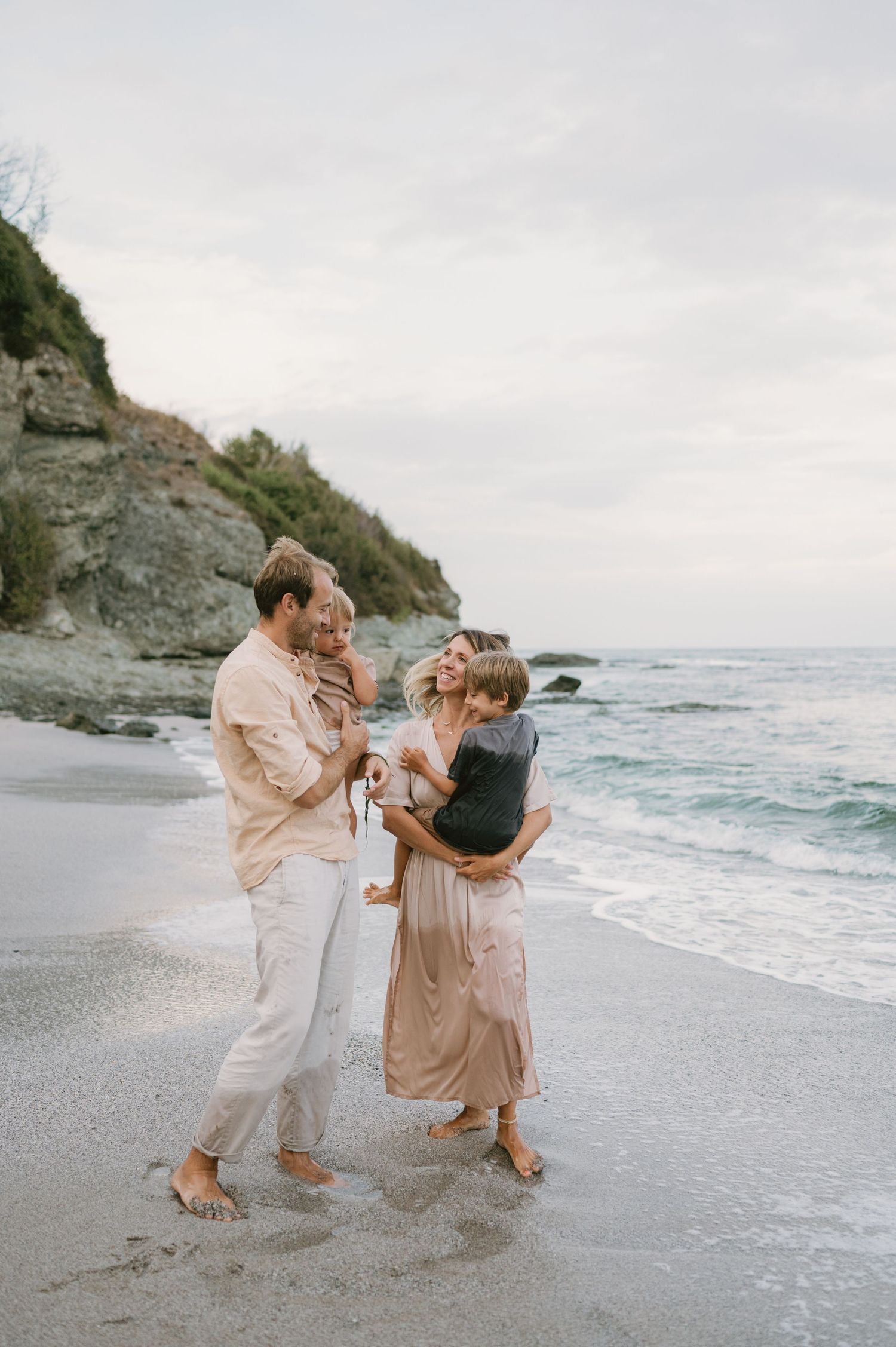 Family of four enjoying a beach walk at sunset, parents holding children with ocean waves and rocky cliffs in background.